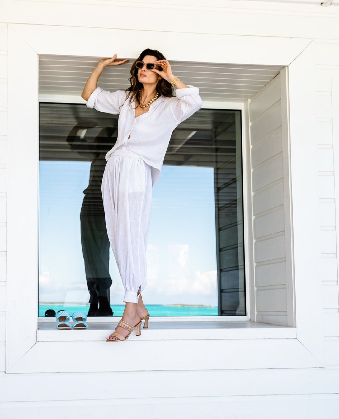 Woman in white blouse and pants standing at a window, wearing sunglasses and high heels, with flip-flops on the windowsill, overlooking an ocean view.