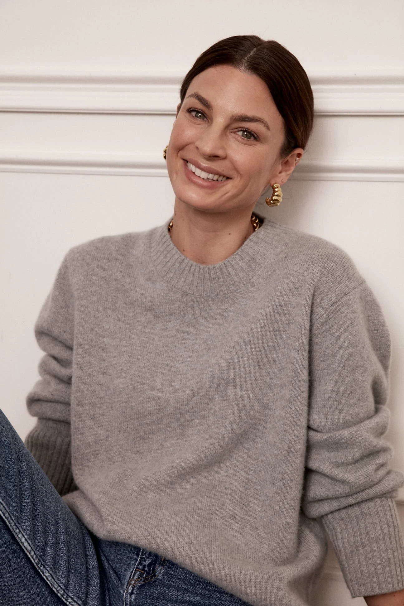 A woman with brown hair wearing gold earrings and a grey sweater, smiling indoors against a white wall.