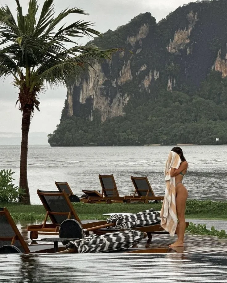 A woman stands on a poolside with a towel over her head, near beach chairs, overlooking water and lush green mountains in the background.