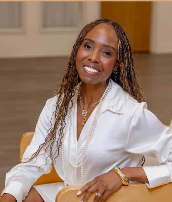 A woman with long hair smiling while sitting at a table; she is wearing a white blouse, layered pearl necklaces, gold jewelry, and is in a well-lit room with wooden flooring and neutral-colored walls.