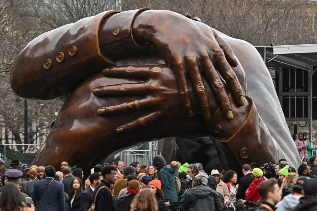 Bronze sculpture of two hands, one large and one small, intertwined and covering an eye, surrounded by a crowd of people outdoors.