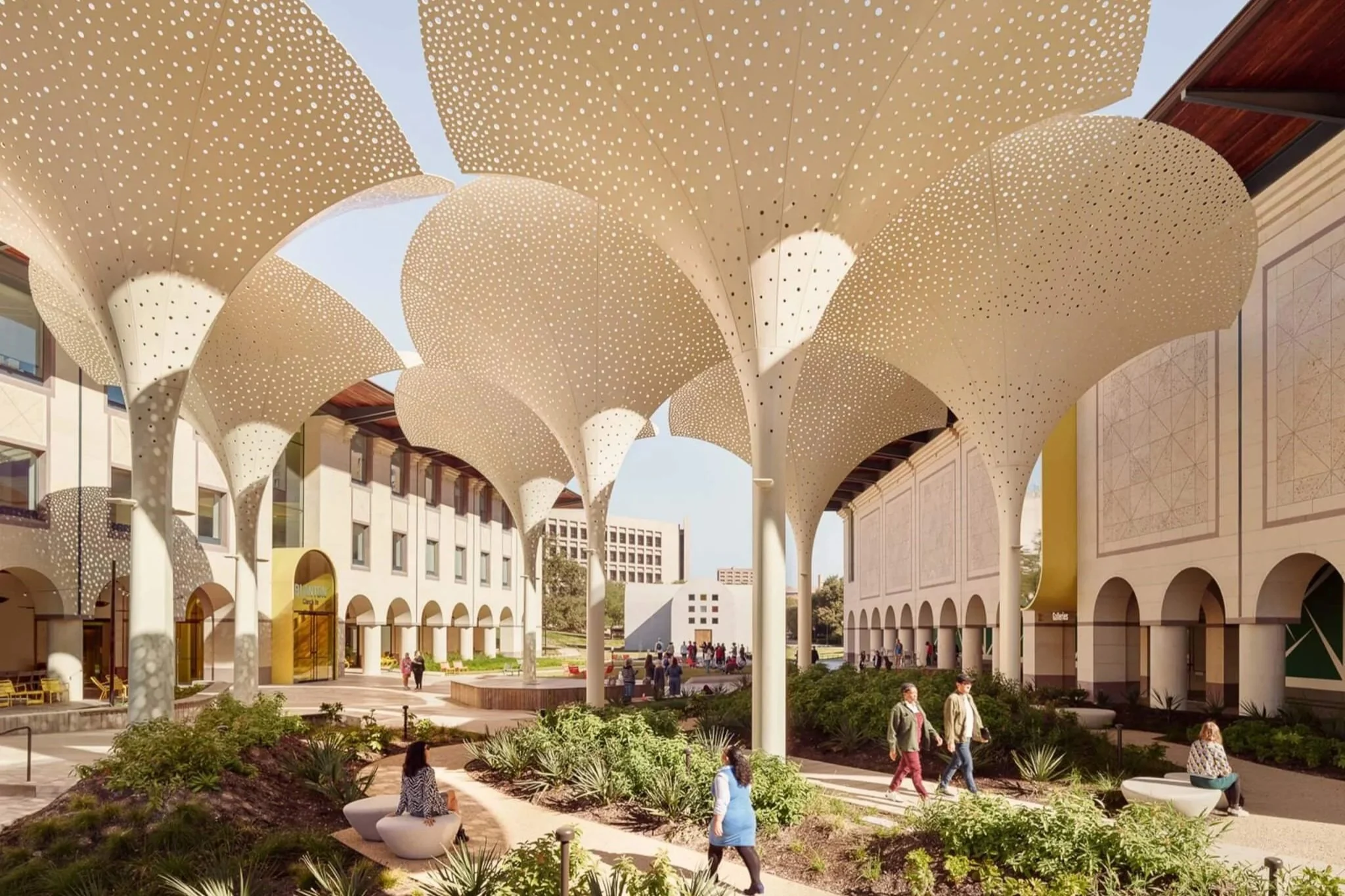 A large outdoor space with modern architecture, featuring distinctive curved, perforated canopy structures overhead, surrounding a landscaped courtyard with benches, plants, and people walking and sitting.