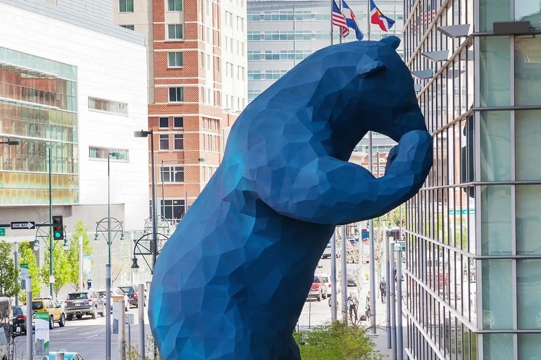 Large blue bear sculpture peering into a glass building in an urban area.