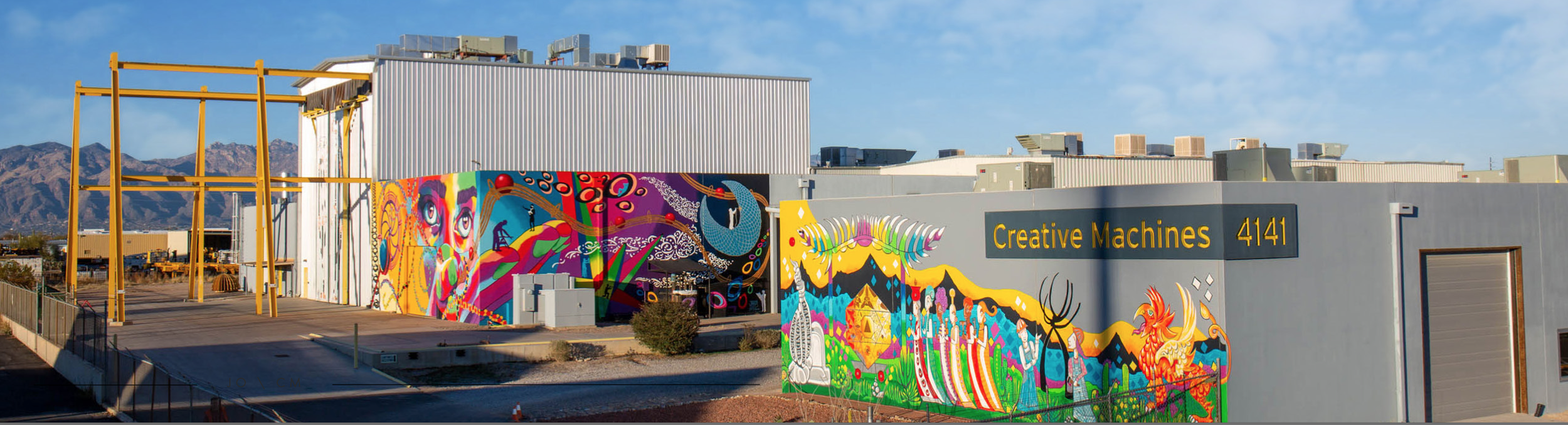 Industrial building with colorful murals and yellow metal framework, mountains in background.