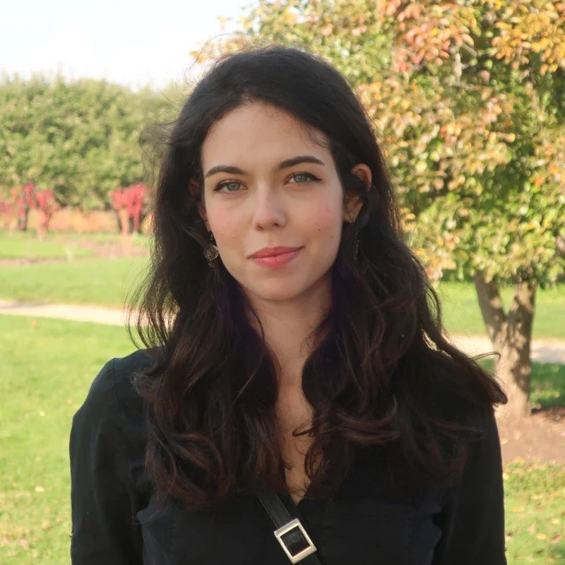 Person with long dark hair wearing a black top, standing outdoors in a park with trees and grass in the background.