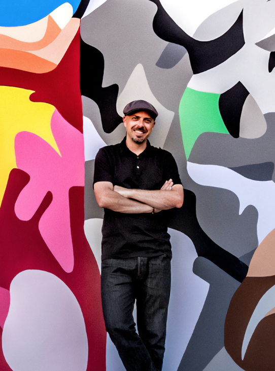A person wearing a flat cap and dark clothing stands in front of a colorful abstract mural, smiling and with arms crossed.