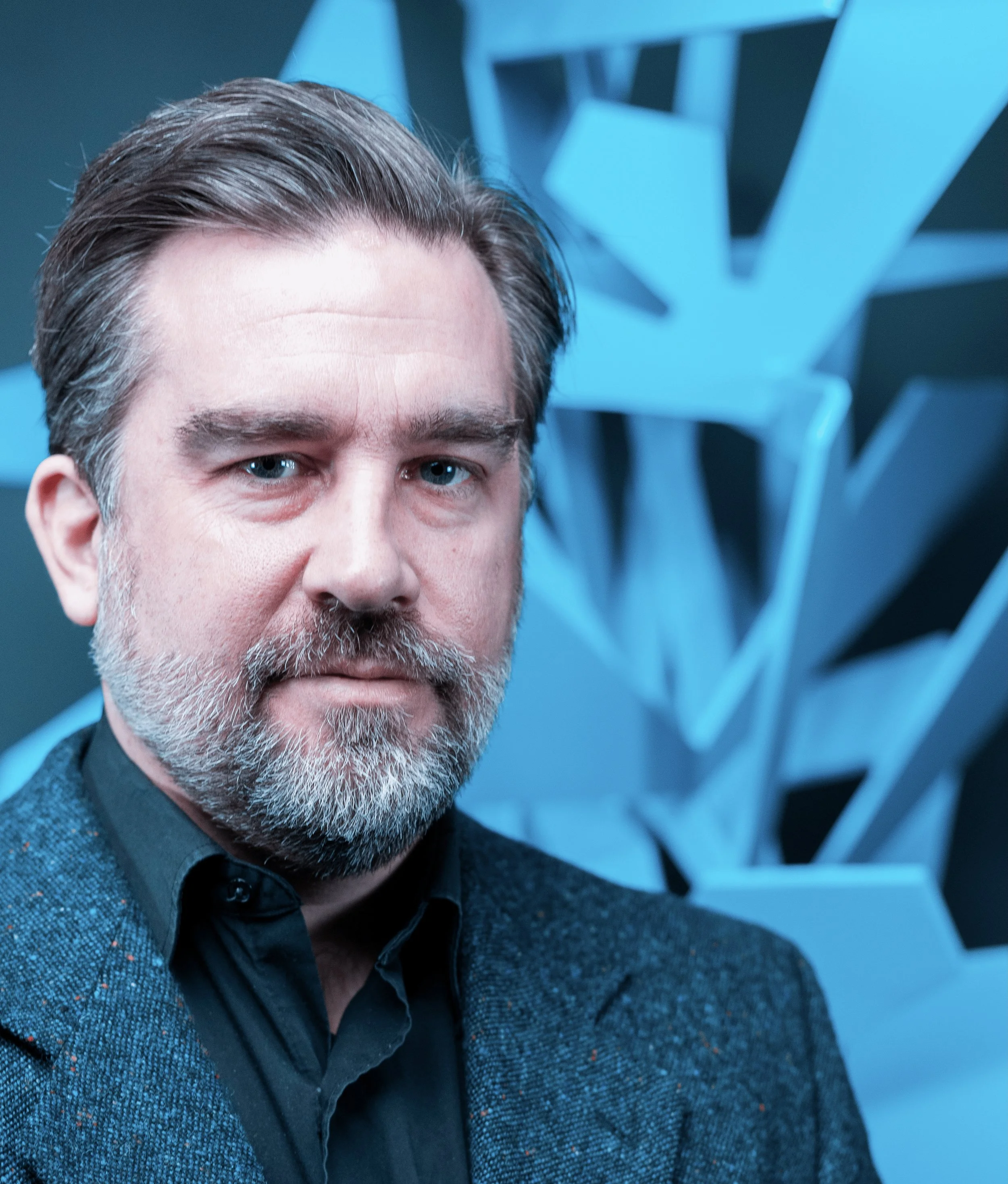 Man with a gray beard and suit in front of a blue abstract background.