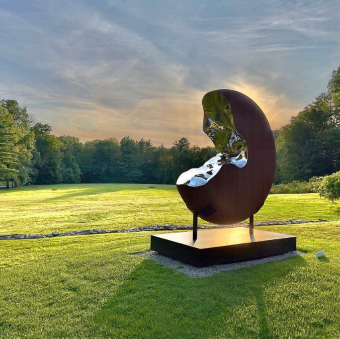 Outdoor sculpture of a reflective metal oval shape on a grassy lawn with trees and a sunrise or sunset in the background.
