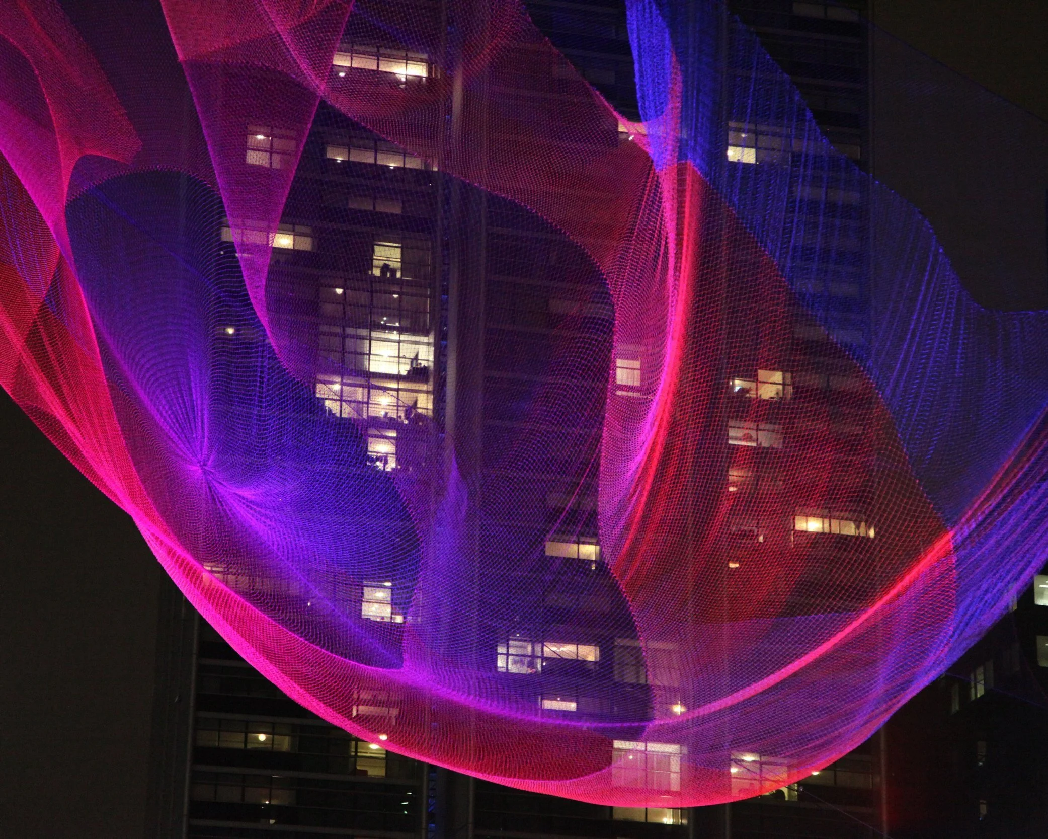Colorful net sculpture illuminated at night in front of a glass building.