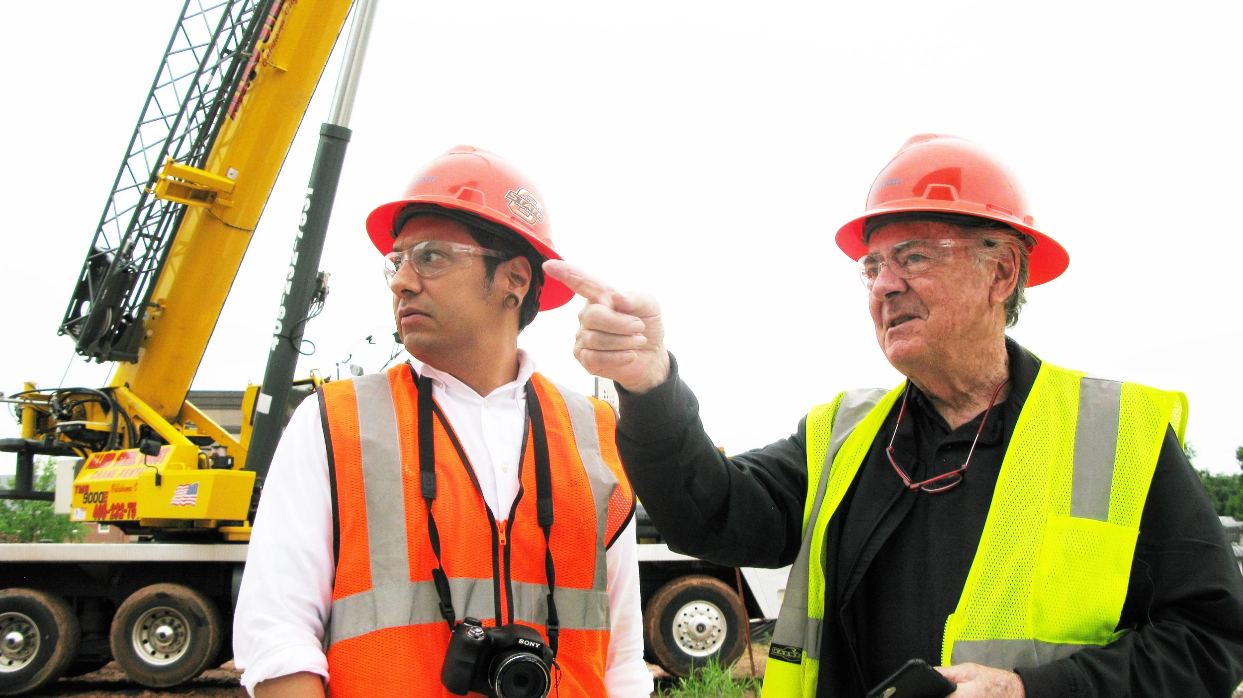 Two construction workers wearing orange safety vests and hard hats stand in front of a yellow crane. One is pointing, while the other looks in the same direction. There is a camera hanging around one worker's neck.