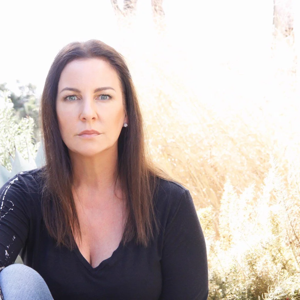 Woman with long dark hair wearing a black top sitting outdoors in sunlight, with dried grass in the background.