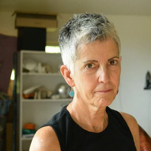 A person with short gray hair wearing a sleeveless black shirt, standing indoors with shelves in the background.