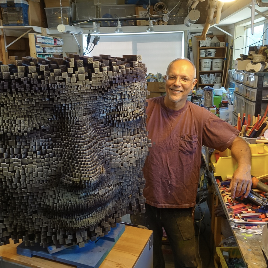 Artist standing next to a large, pixelated face sculpture made from wooden blocks in a workshop filled with tools and supplies.