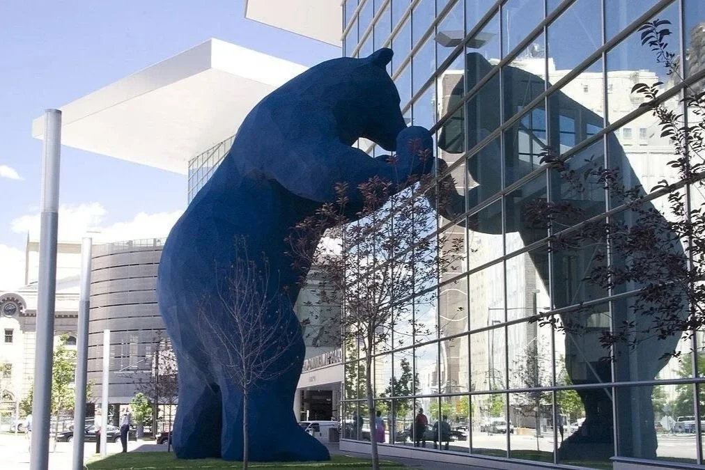 A large blue bear sculpture is reflected in the glass of a modern building, with trees and people in the background.
