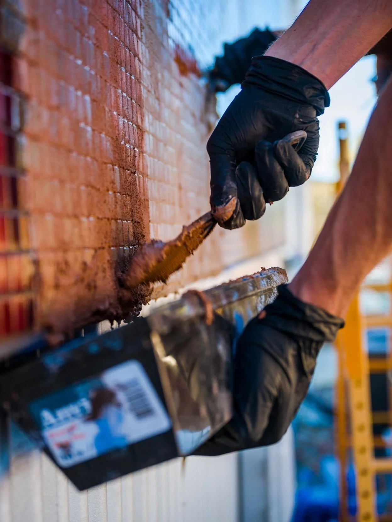 Close-up of a person wearing black gloves applying mortar to a tiled surface using a trowel.