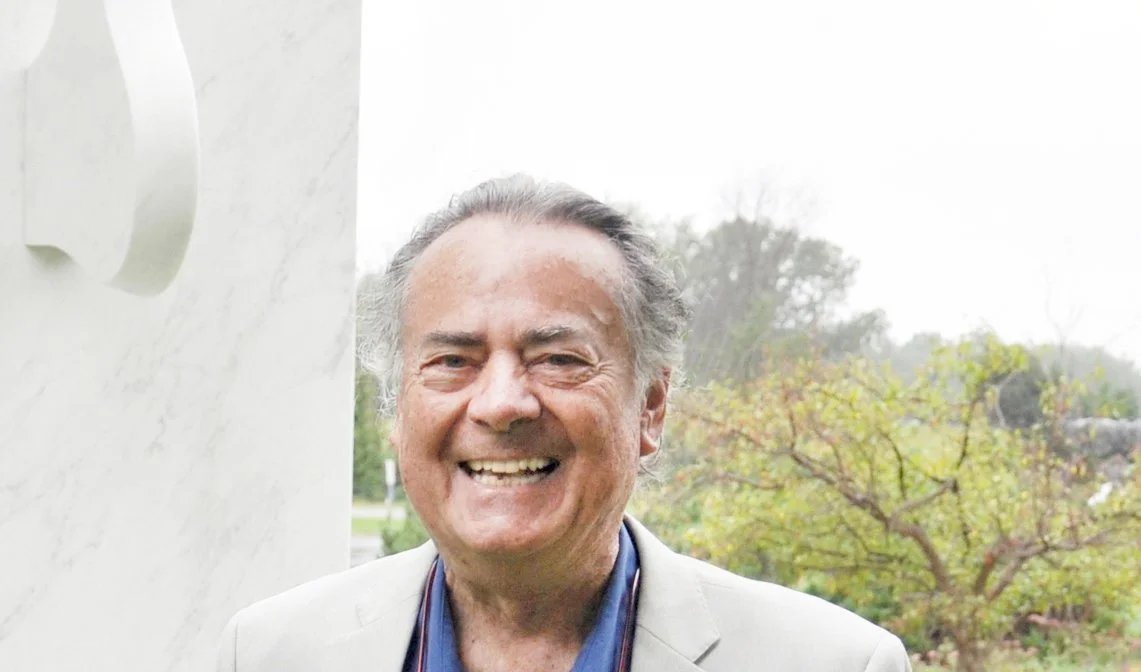 Smiling man wearing a light-colored blazer, standing outdoors with trees in the background.
