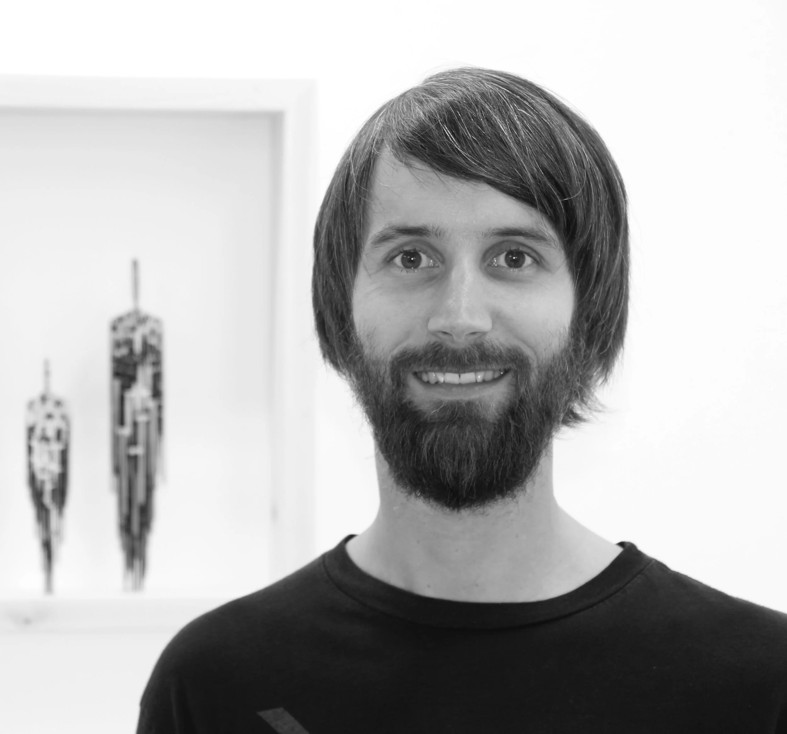 Black and white photo of a smiling man with a beard, standing indoors next to wall art.