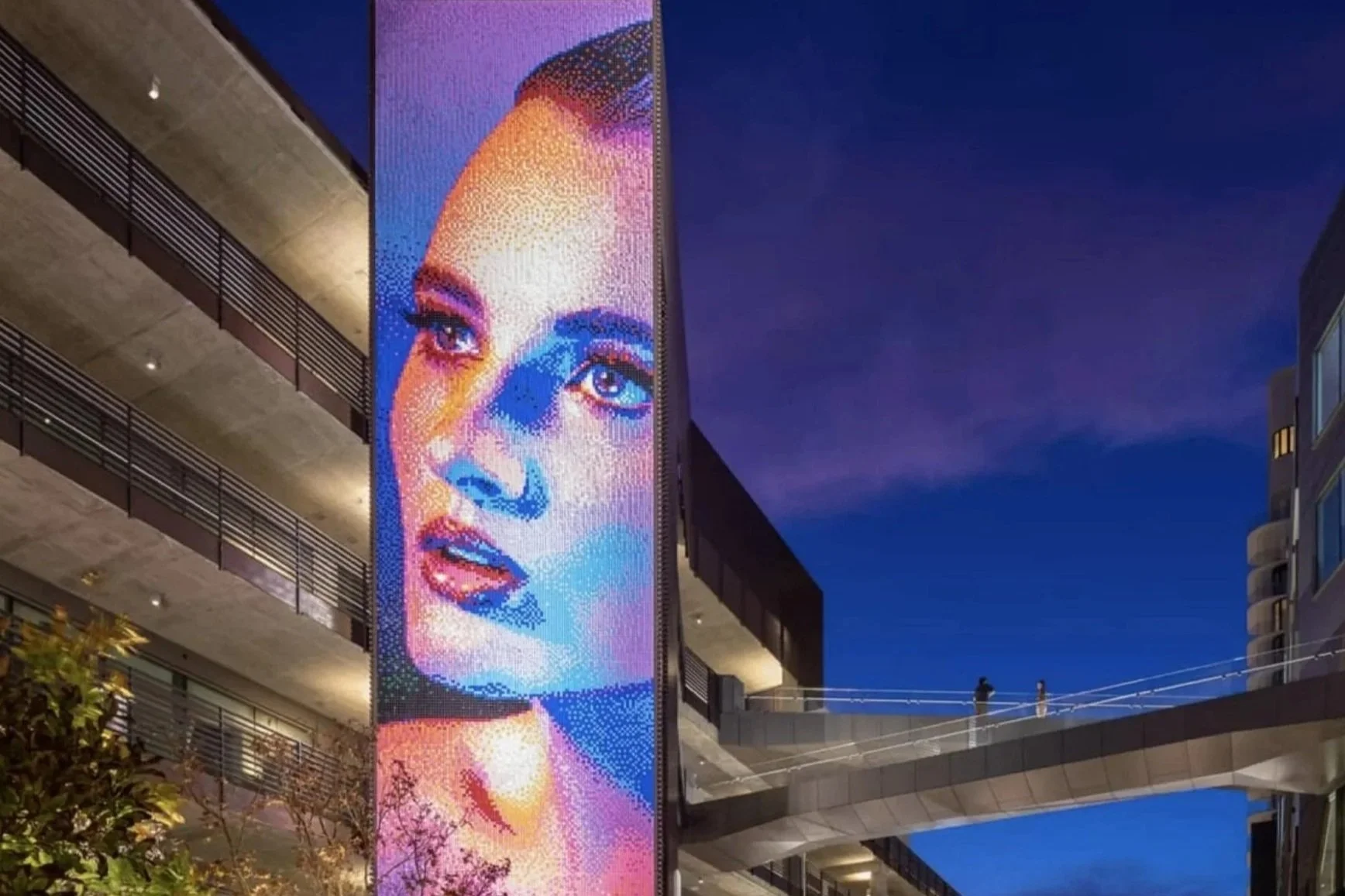 Nighttime cityscape featuring a large vertical electronic billboard displaying a colorful portrait of a woman's face with blue, pink, and purple tones. The billboard is attached to a modern building with balconies and railings, and there is a skybridge with two people walking on it connecting buildings. The sky is dark blue with some light clouds.