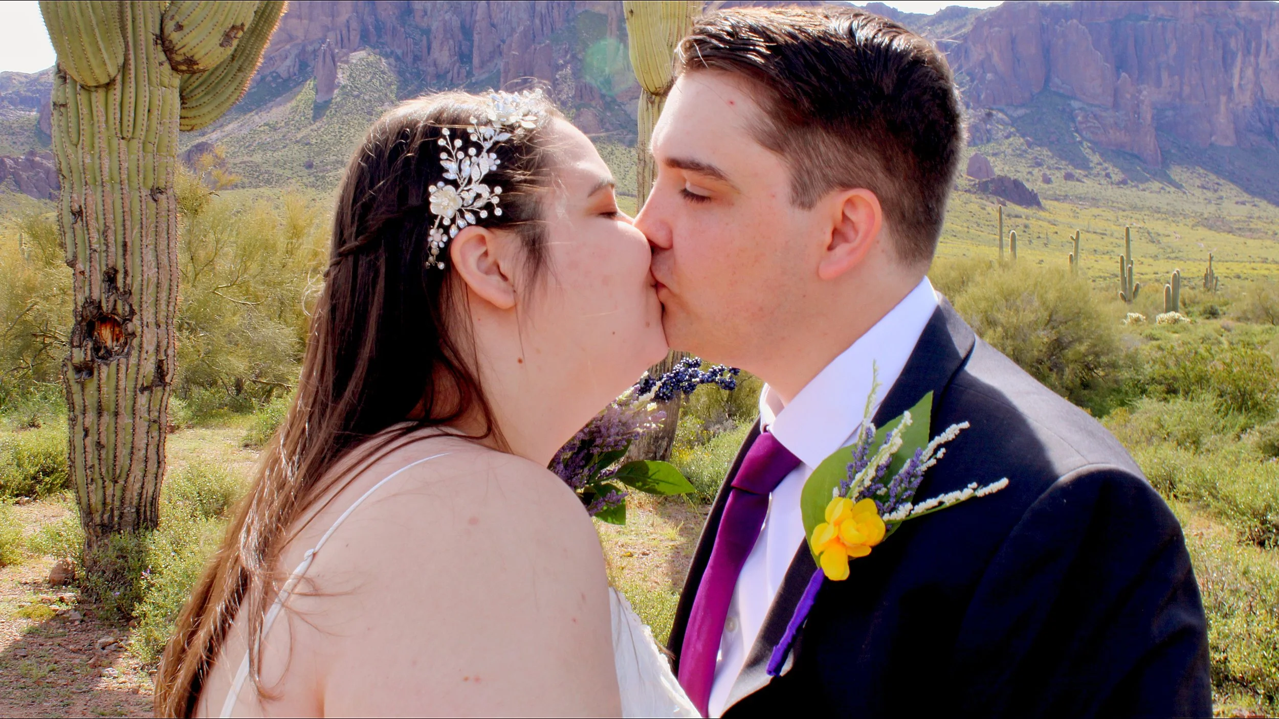 A bride and groom kissing in a desert landscape with cacti and mountains in the background.