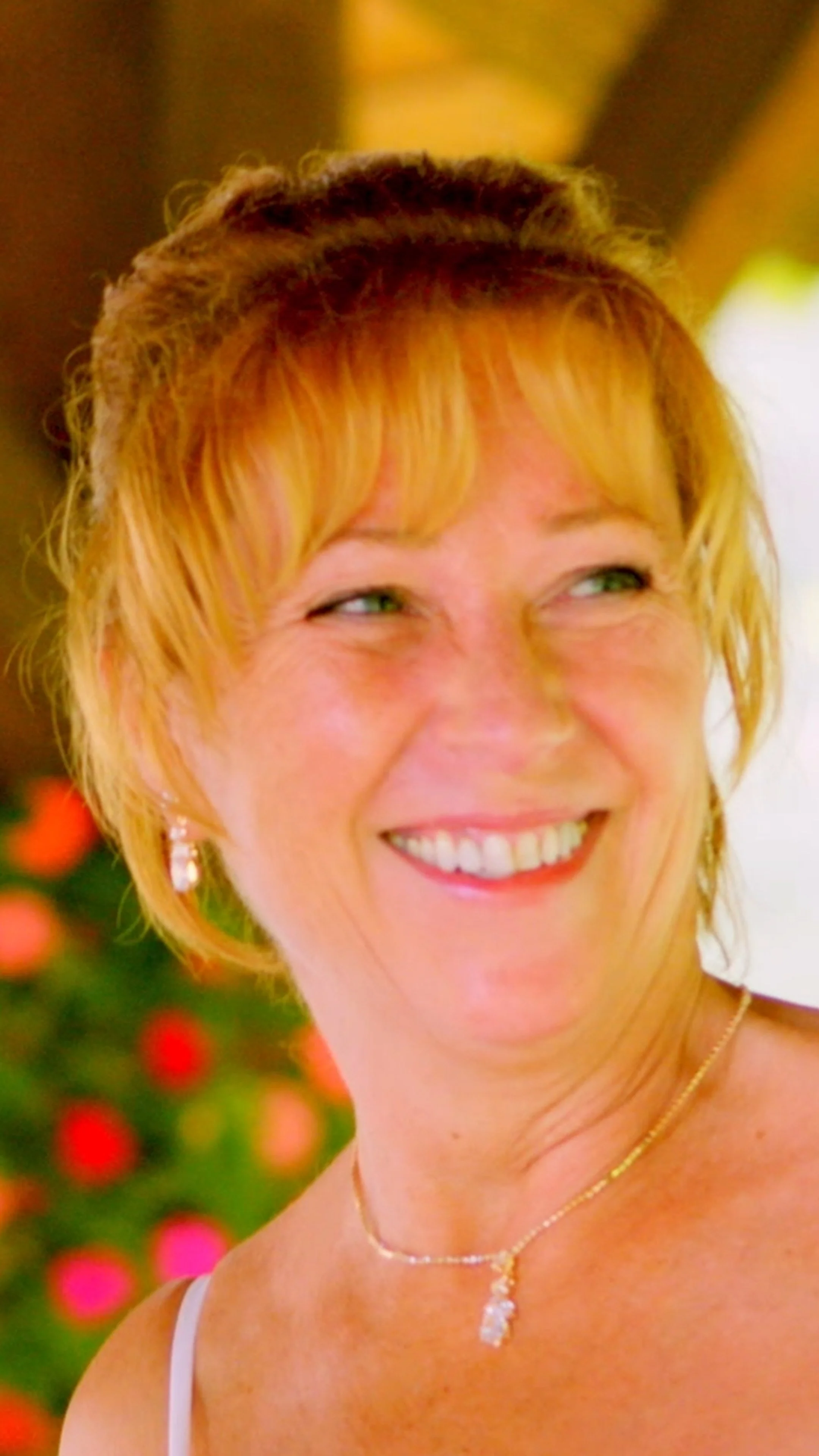 A woman with short blonde hair smiling, wearing jewelry, with a decorated Christmas tree in the background.