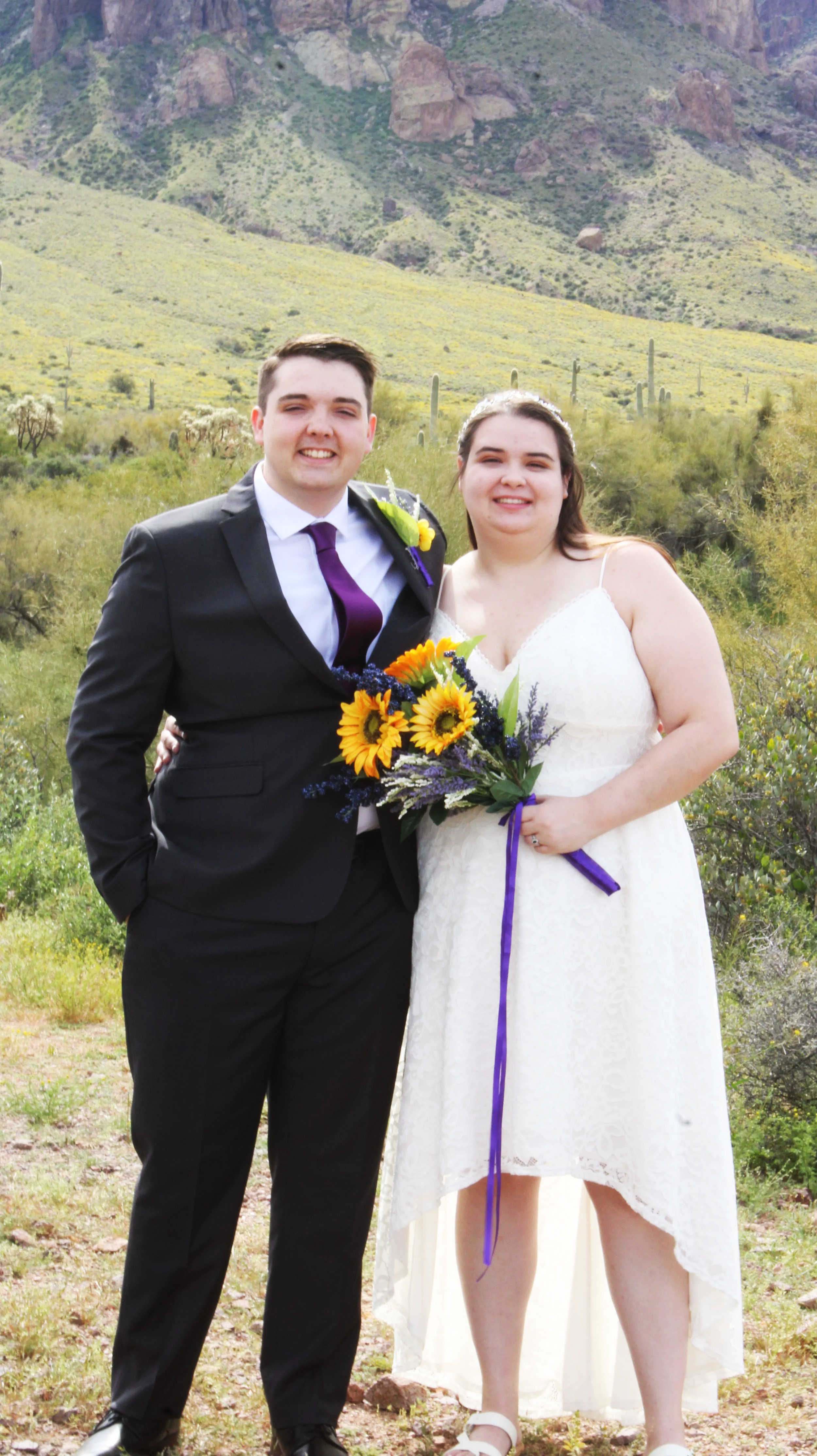 A wedding couple standing outdoors in a desert landscape with mountains and cacti in the background. The groom wears a black suit with a white shirt and purple tie, and the bride wears a white lace dress holding a bouquet of sunflowers and purple flo