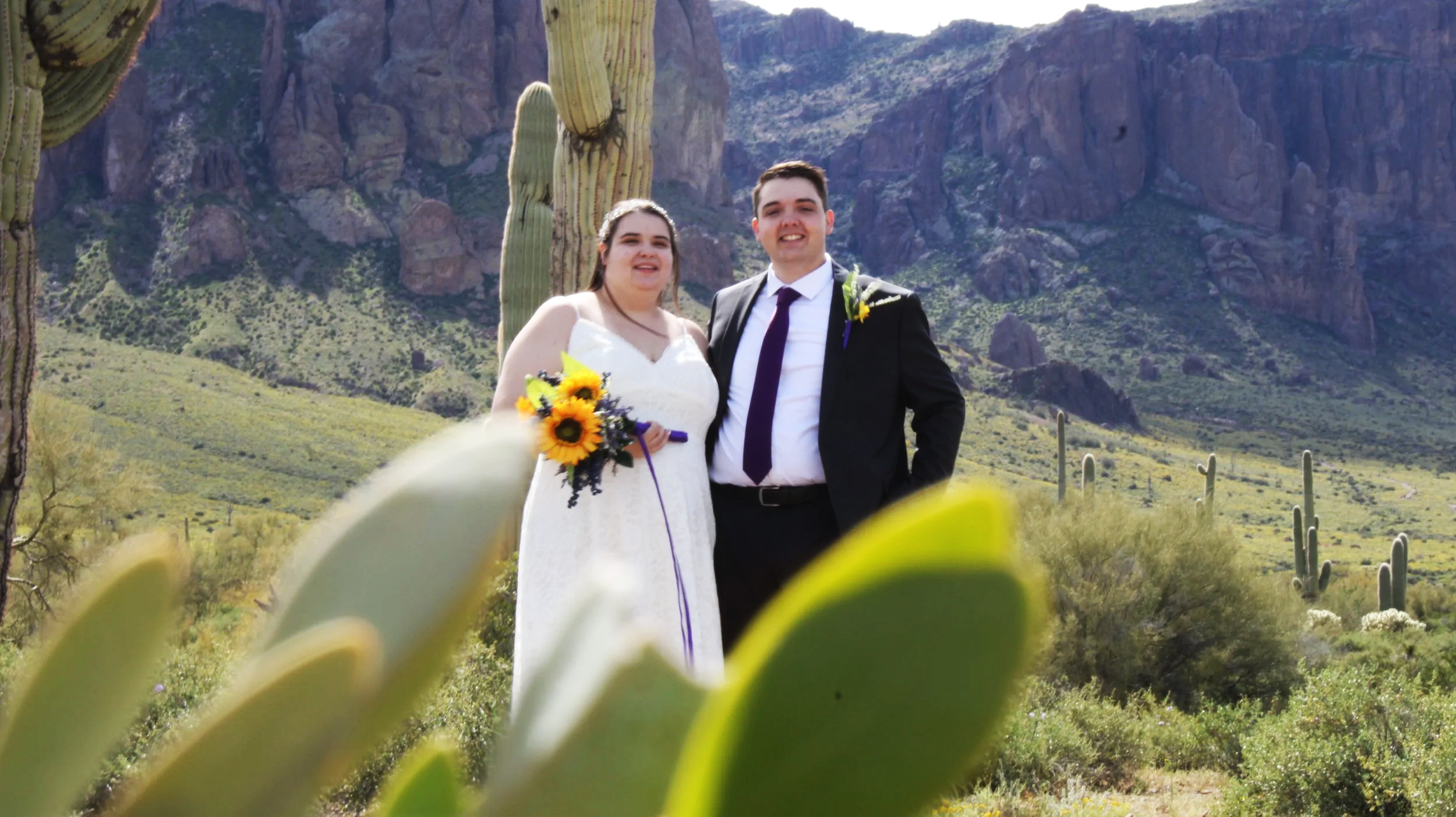 A bride and groom standing together outdoors in a desert landscape with cacti, mountains, and blue sky in the background.