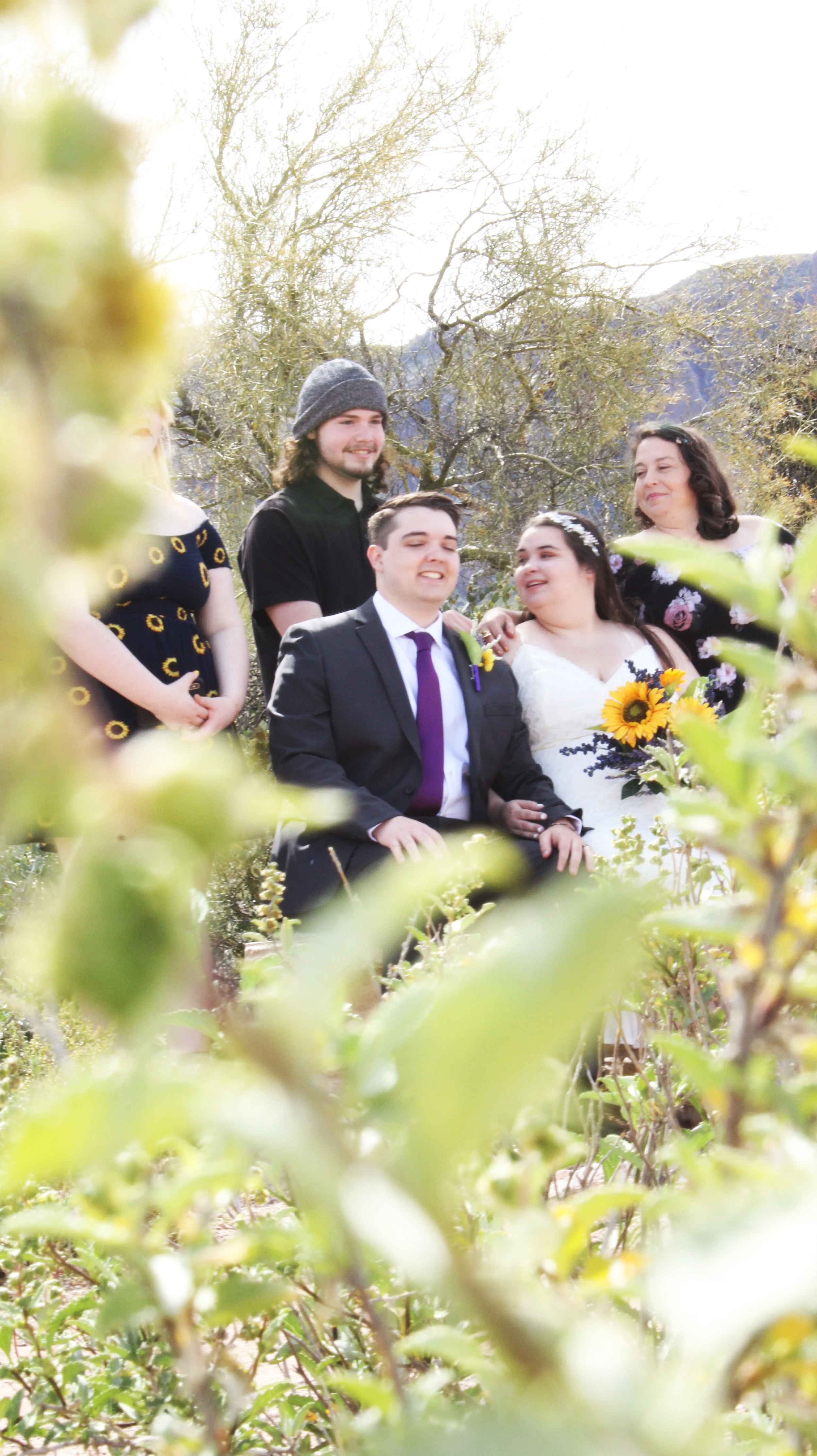 A wedding group photo outdoors with five people, including a bride and groom, standing and sitting among green foliage with trees and mountains in the background.