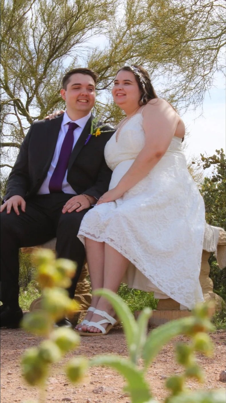 A newlywed couple sitting outdoors on a stone bench under a large tree, smiling. The bride is in a white lace dress with a floral headpiece, and the groom is in a black suit with a purple tie and a yellow boutonniere.