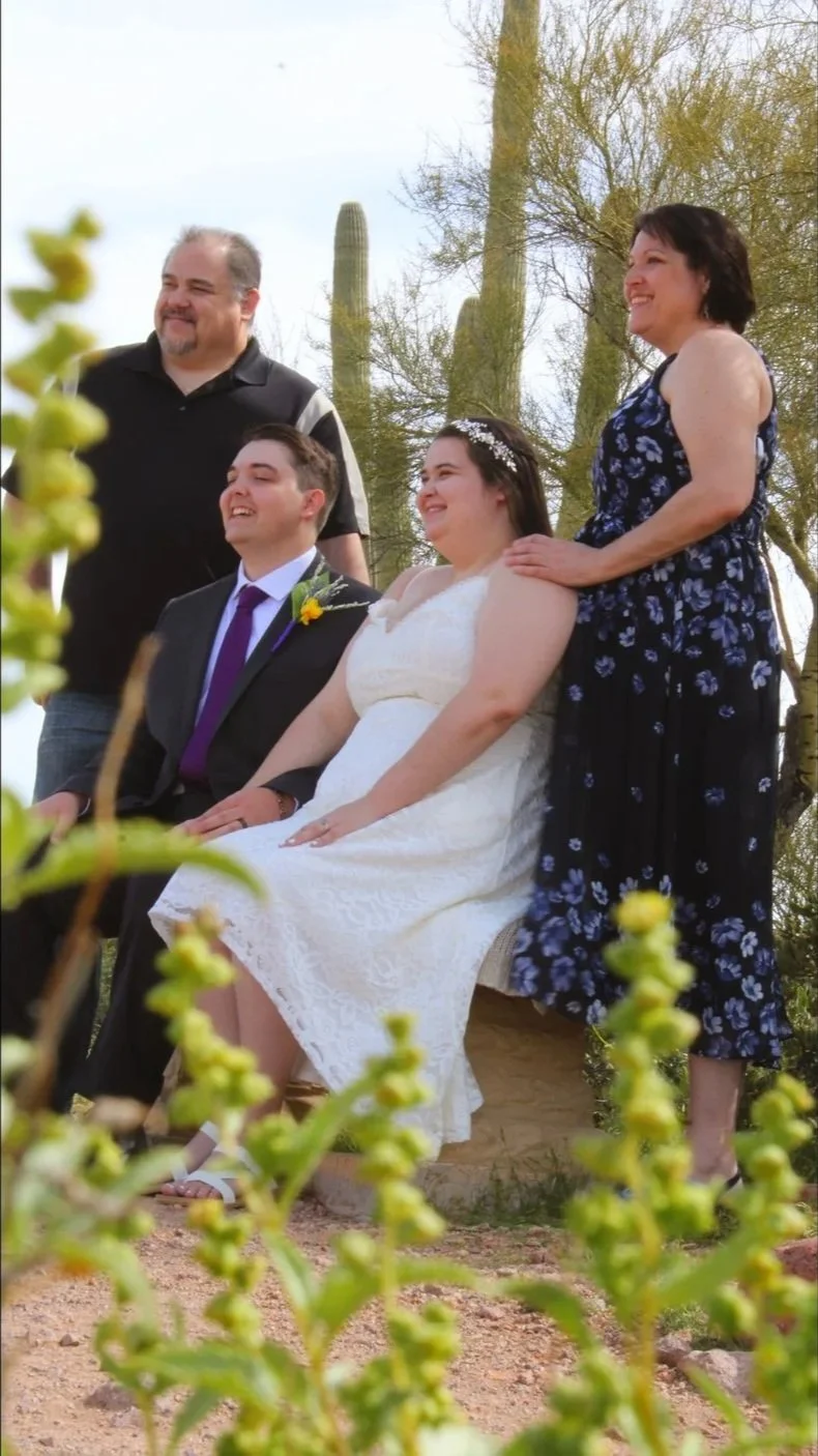 A group of four people outdoors during a wedding celebration, with two women and two men smiling and posing for the camera. There are desert trees and cacti in the background.