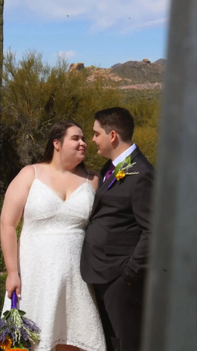 A bride and groom standing close together outdoors, with a desert landscape and mountains in the background. The bride is in a white lace dress holding a bouquet of flowers, and the groom is in a black suit with a boutonniere. They are looking into e