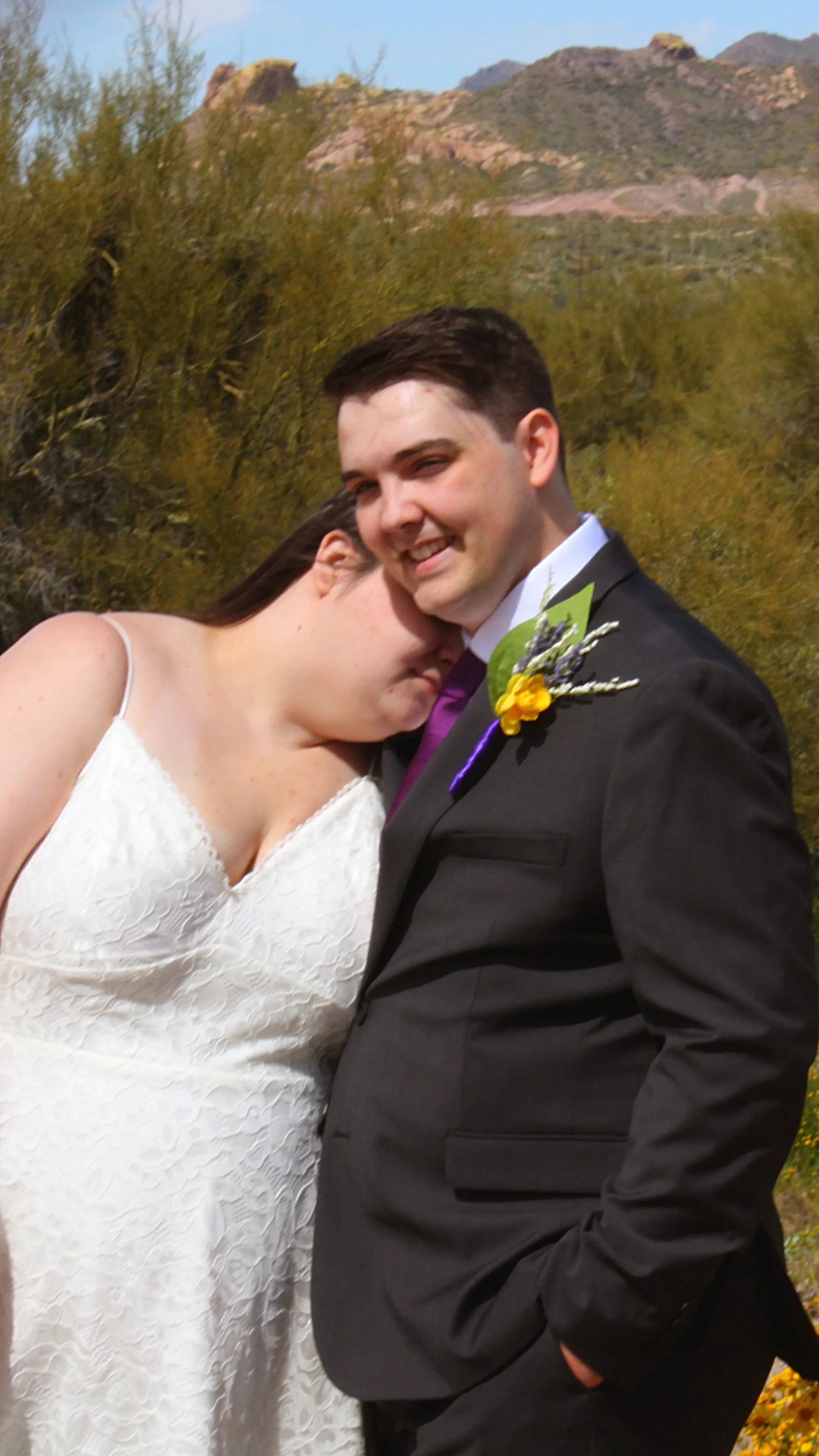 A bride and groom at their wedding outdoors, with desert mountains and trees in the background. The bride is leaning her head on the groom's shoulder, and they are both smiling.