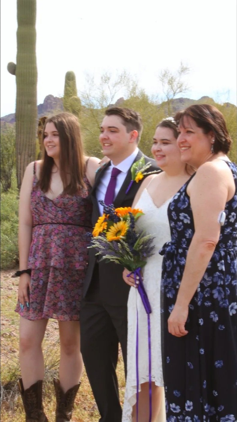Four people standing outdoors, two women and one man dressed in a suit, and one young woman in a white dress holding a bouquet of flowers, with desert plants and mountains in the background.