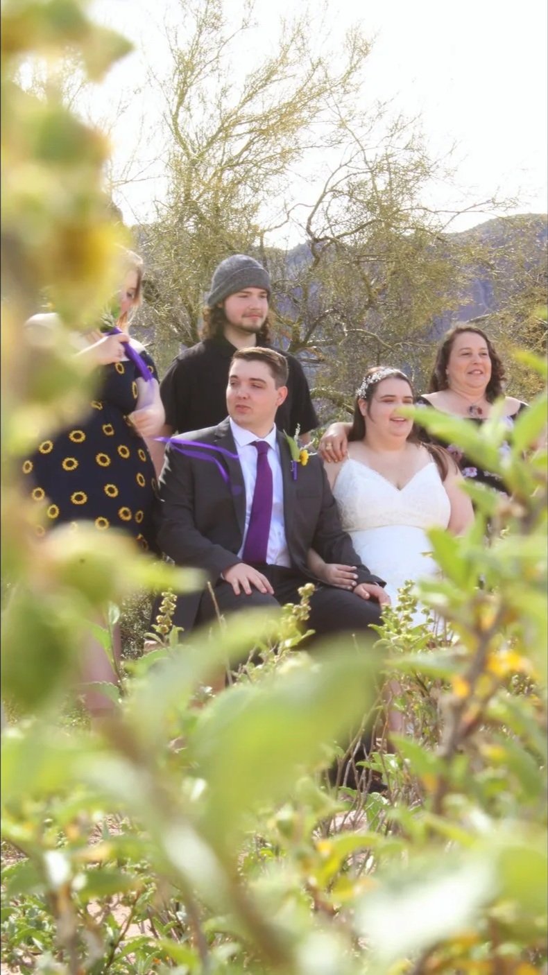 Group of five people outdoors, two women and three men, celebrating a special occasion like a wedding, with a bride in a white dress and a groom in a black suit with a purple tie sitting among the group. Green foliage in the foreground, trees and hil