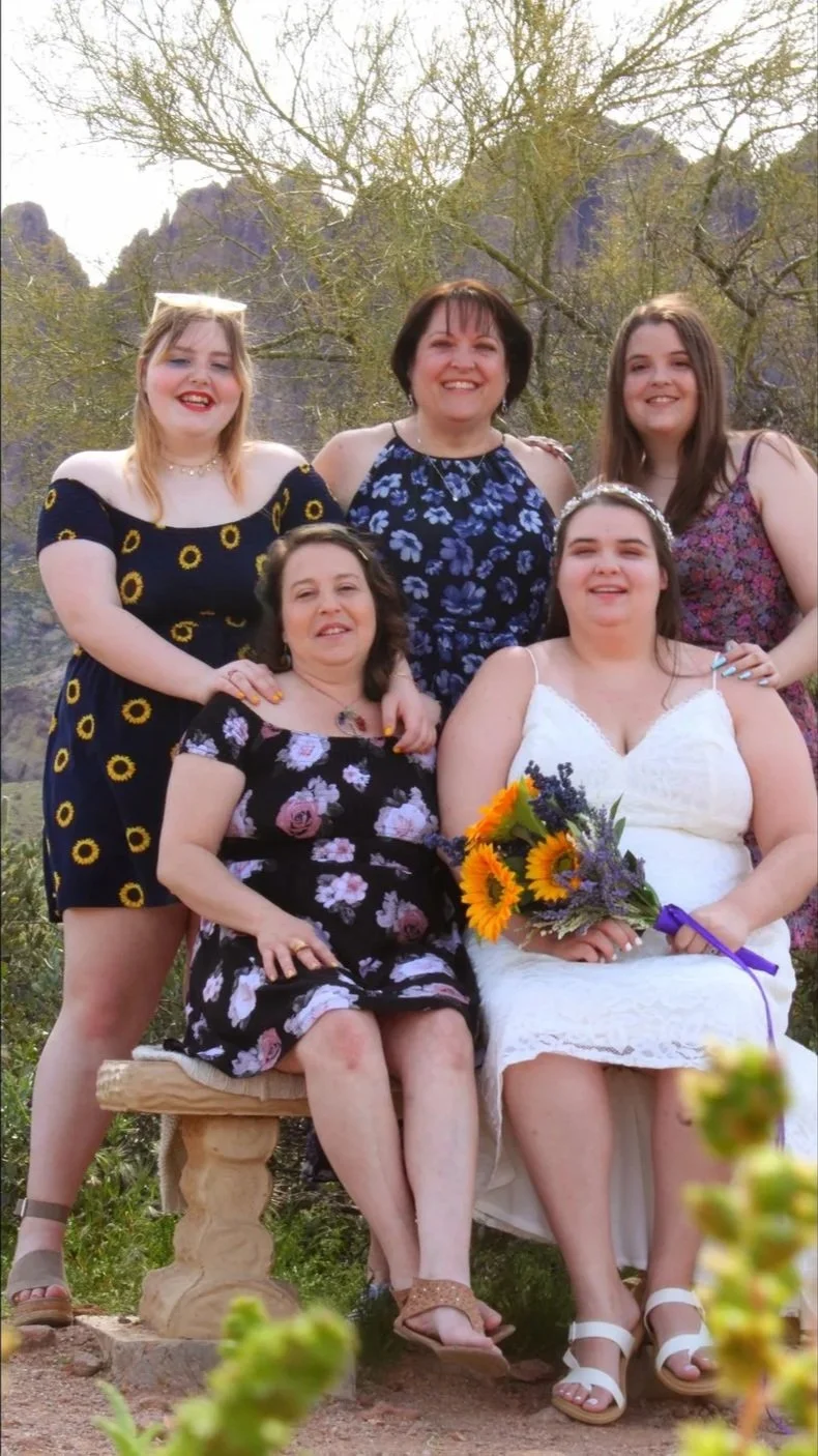 Group of six women, one in a wedding dress holding a bouquet of sunflowers and lavender, posing outdoors during daytime with mountains and trees in background.