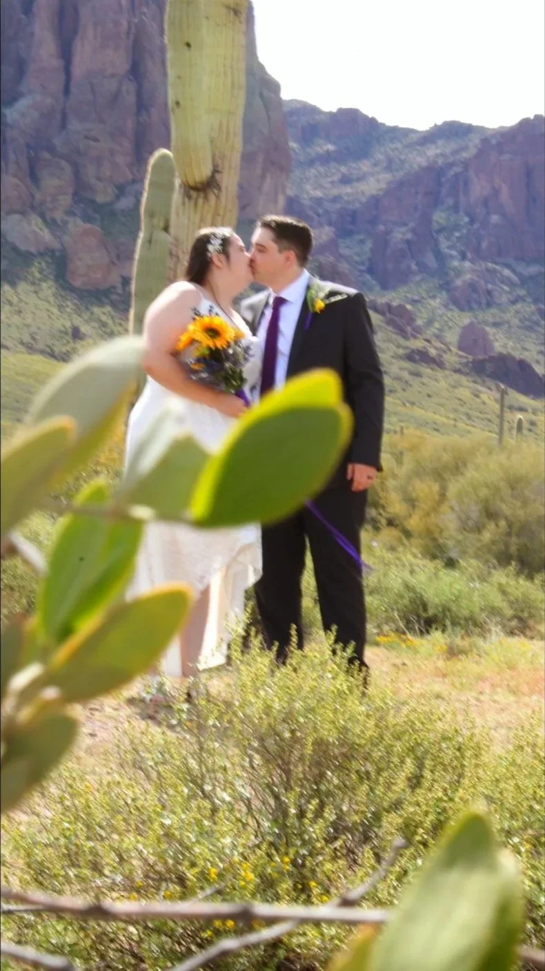 A bride and groom sharing a kiss outdoors in a desert landscape with cacti and mountains in the background.