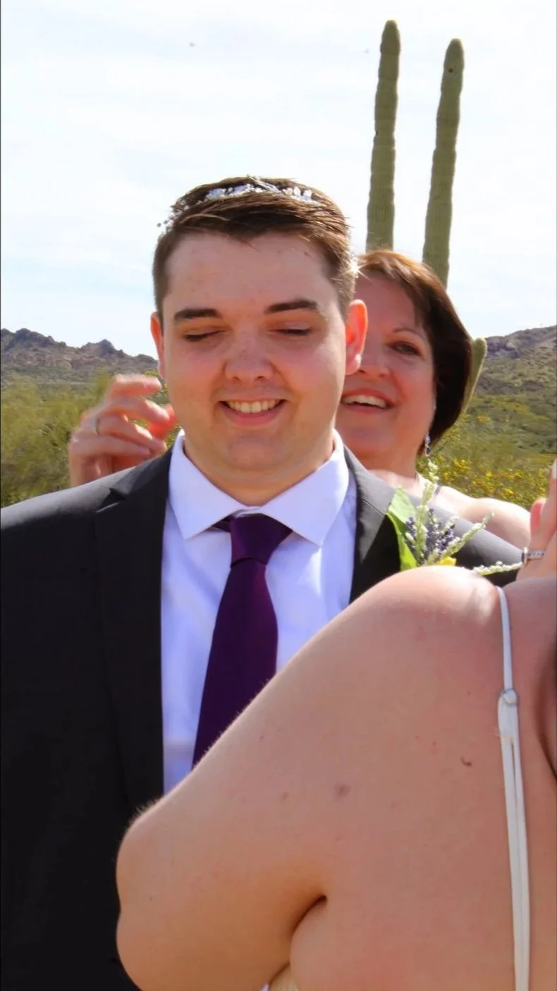 A man in a suit and purple tie smiling outdoors, with a woman and cacti in the background, in a desert landscape.