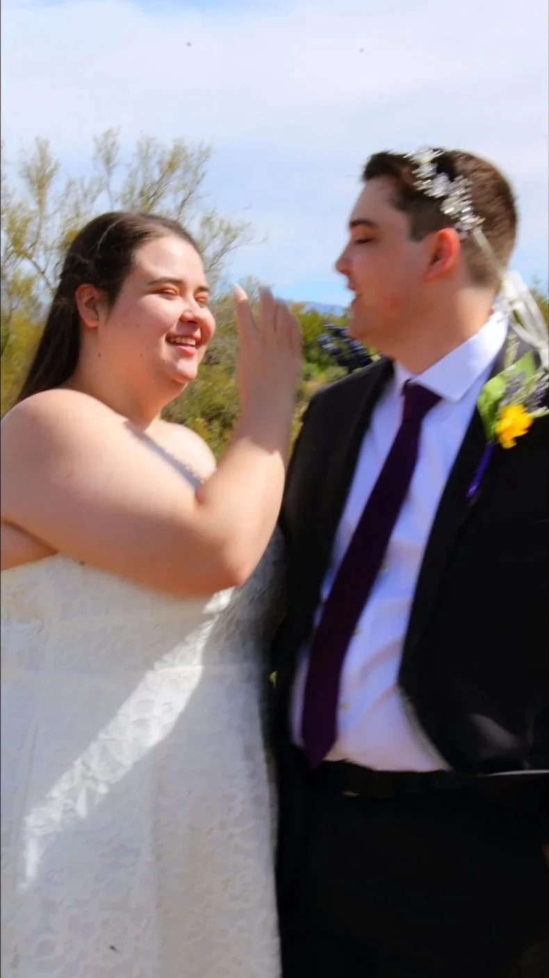 A smiling bride and groom sharing a joyful moment outdoors on their wedding day, with trees and a partly cloudy sky in the background.