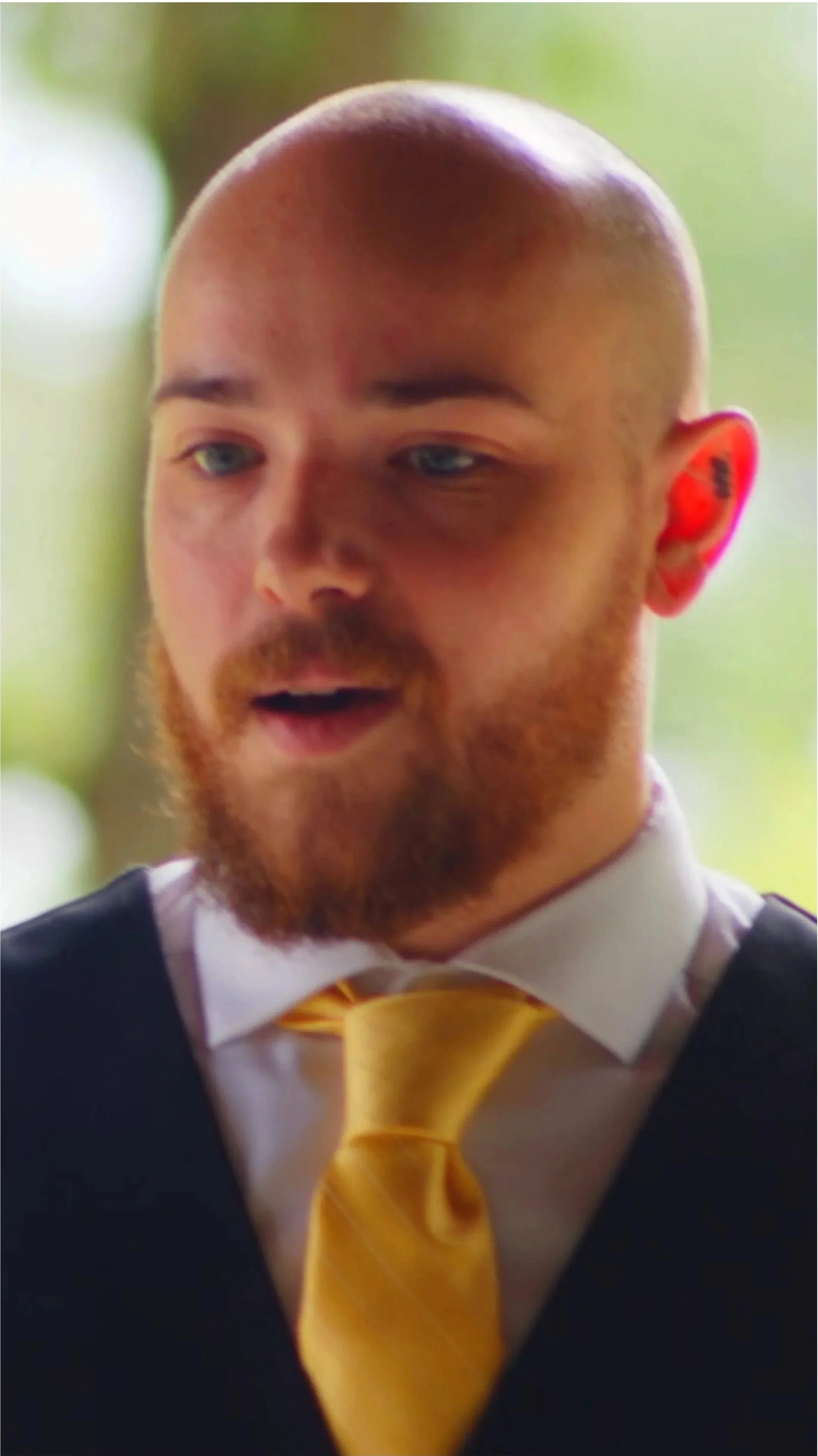 A man with a beard and a shaved head wearing a black vest, white shirt, and yellow tie, looking down with a surprised or interested expression.