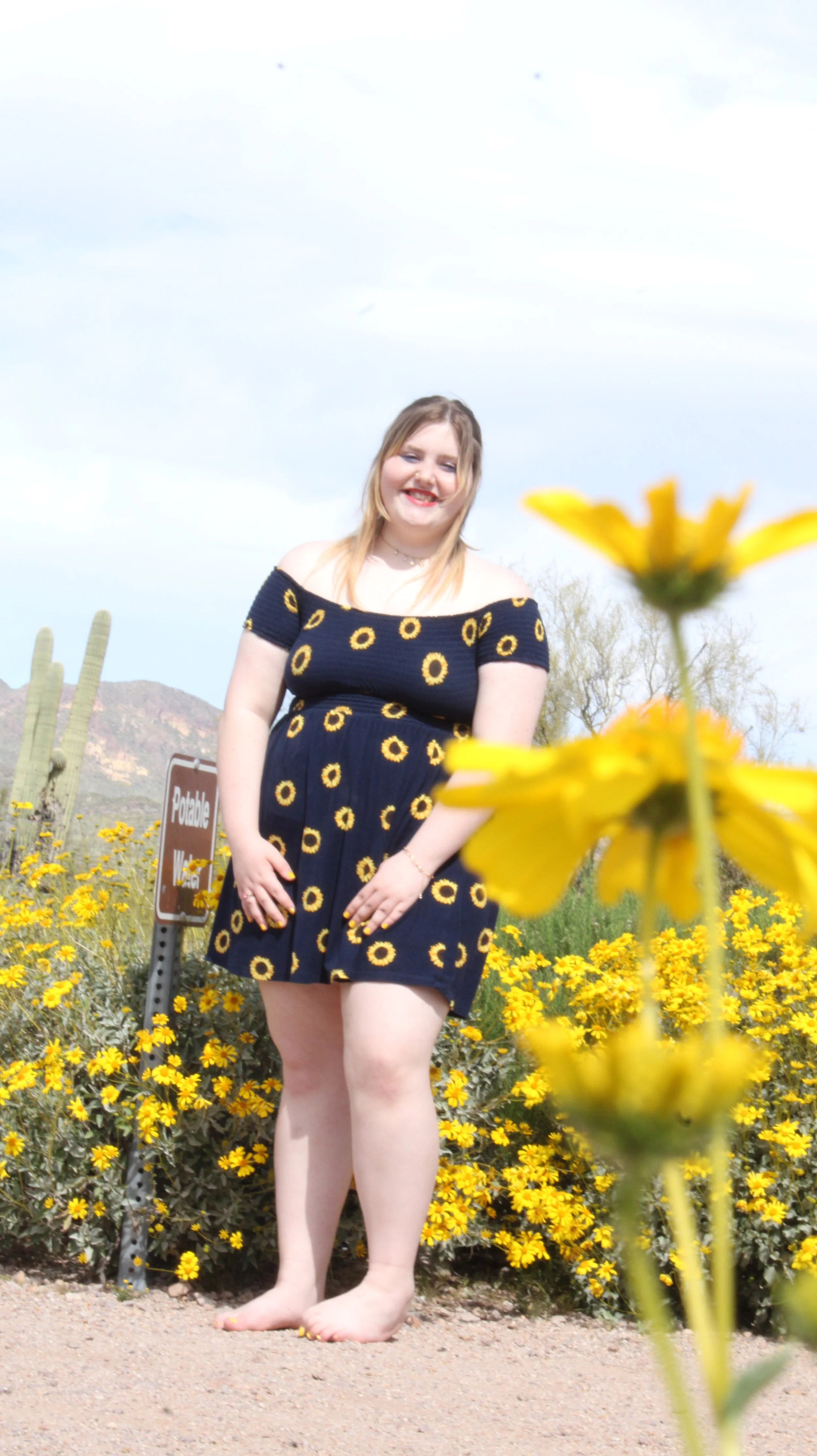 Woman standing barefoot among yellow flowers, wearing a navy dress with sunflower print, smiling outdoors in a desert landscape.