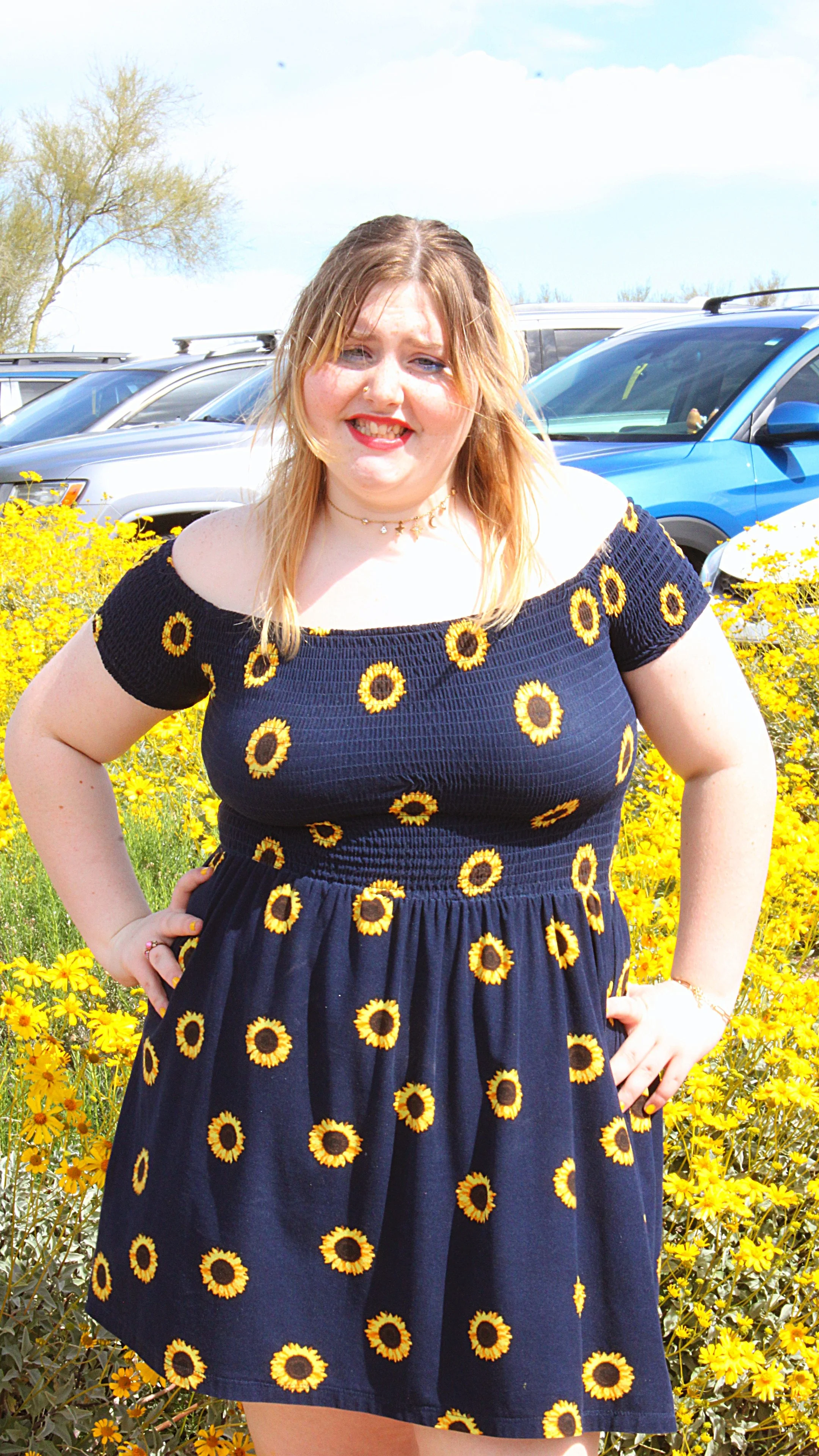 A woman standing outdoors in front of yellow flowers, wearing a navy dress with sunflower patterns, smiling with her hands on her hips.