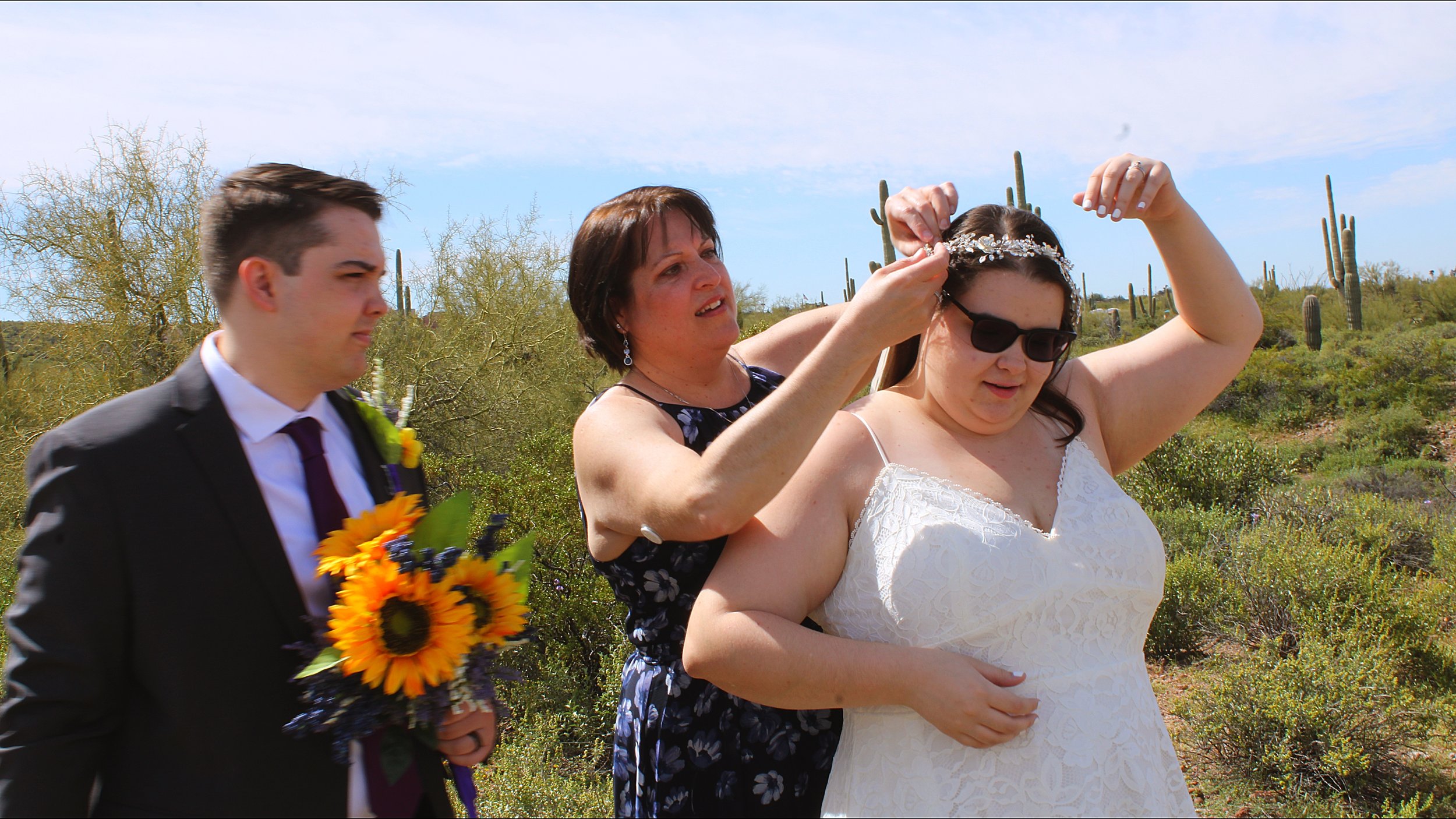 A woman in a white dress is having a crown placed on her head during a wedding ceremony outdoors in a desert area with cacti, while a man in a suit holding a bouquet of sunflowers watches nearby.