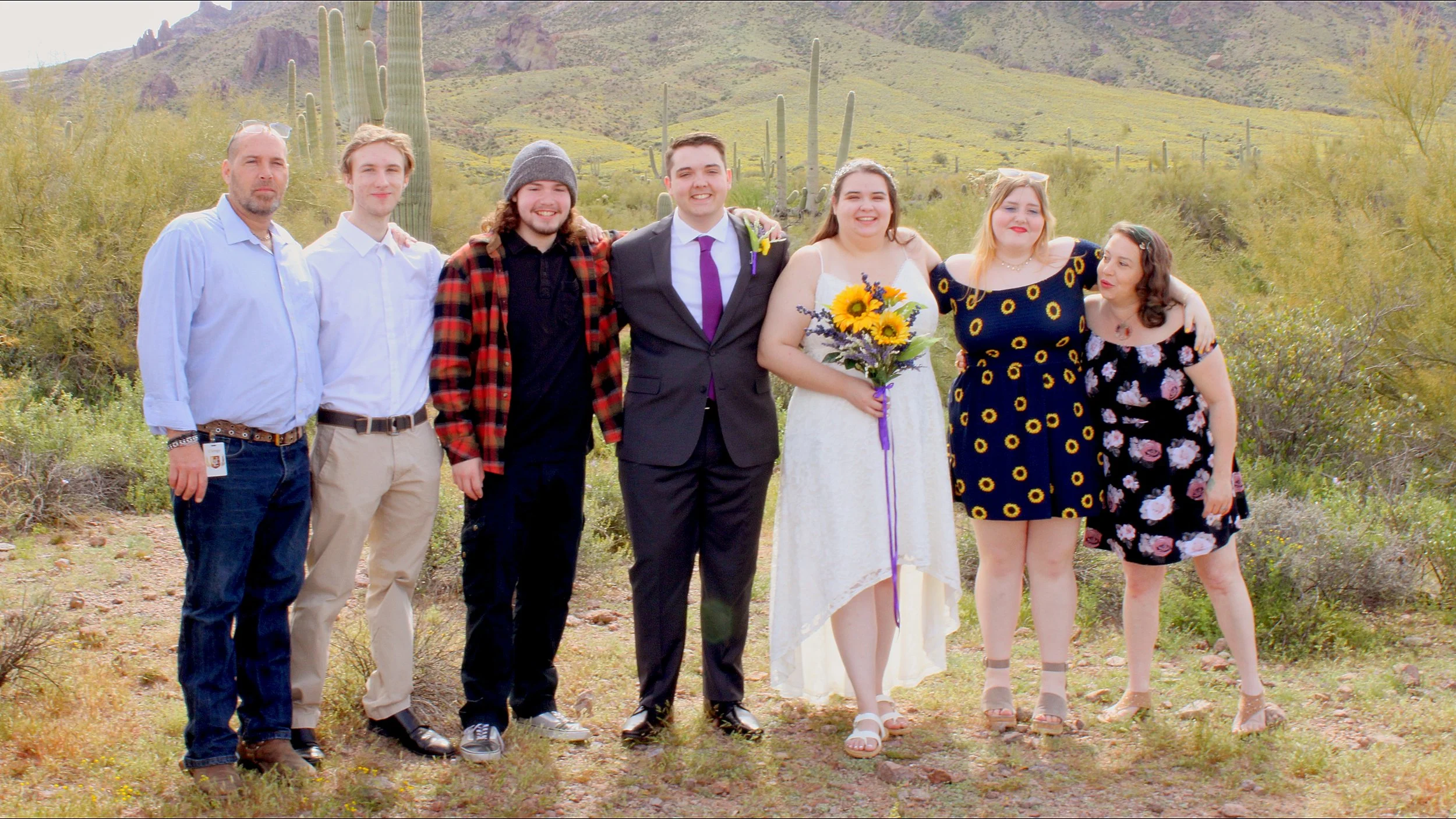Group of seven people standing together outdoors in a desert landscape with tall cacti and hills in the background, dressed in casual and formal clothing.