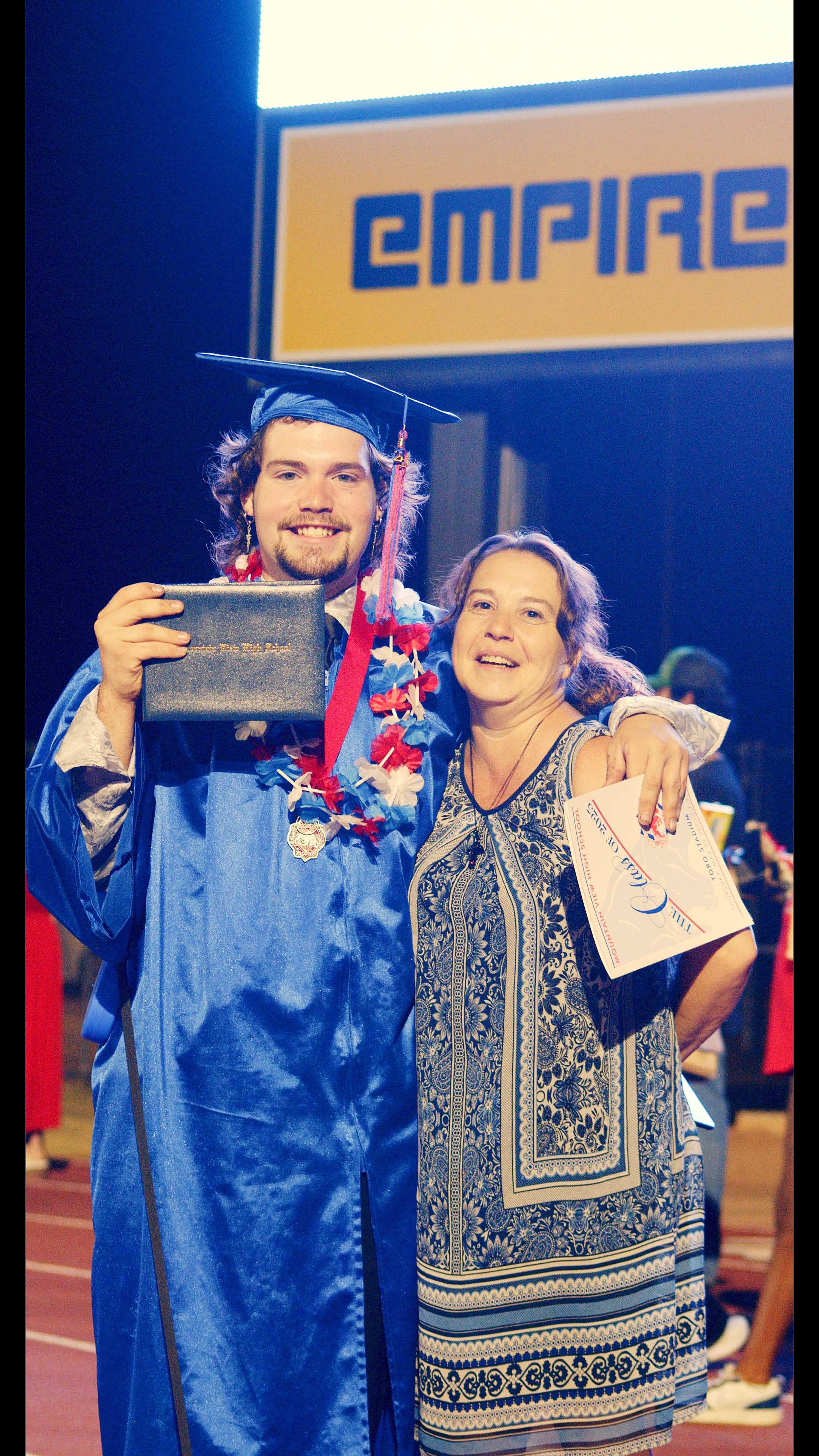 A young man in a blue graduation gown and cap, wearing a lei,, holding a diploma, hugging a woman in a patterned dress, on a stage at a graduation ceremony.