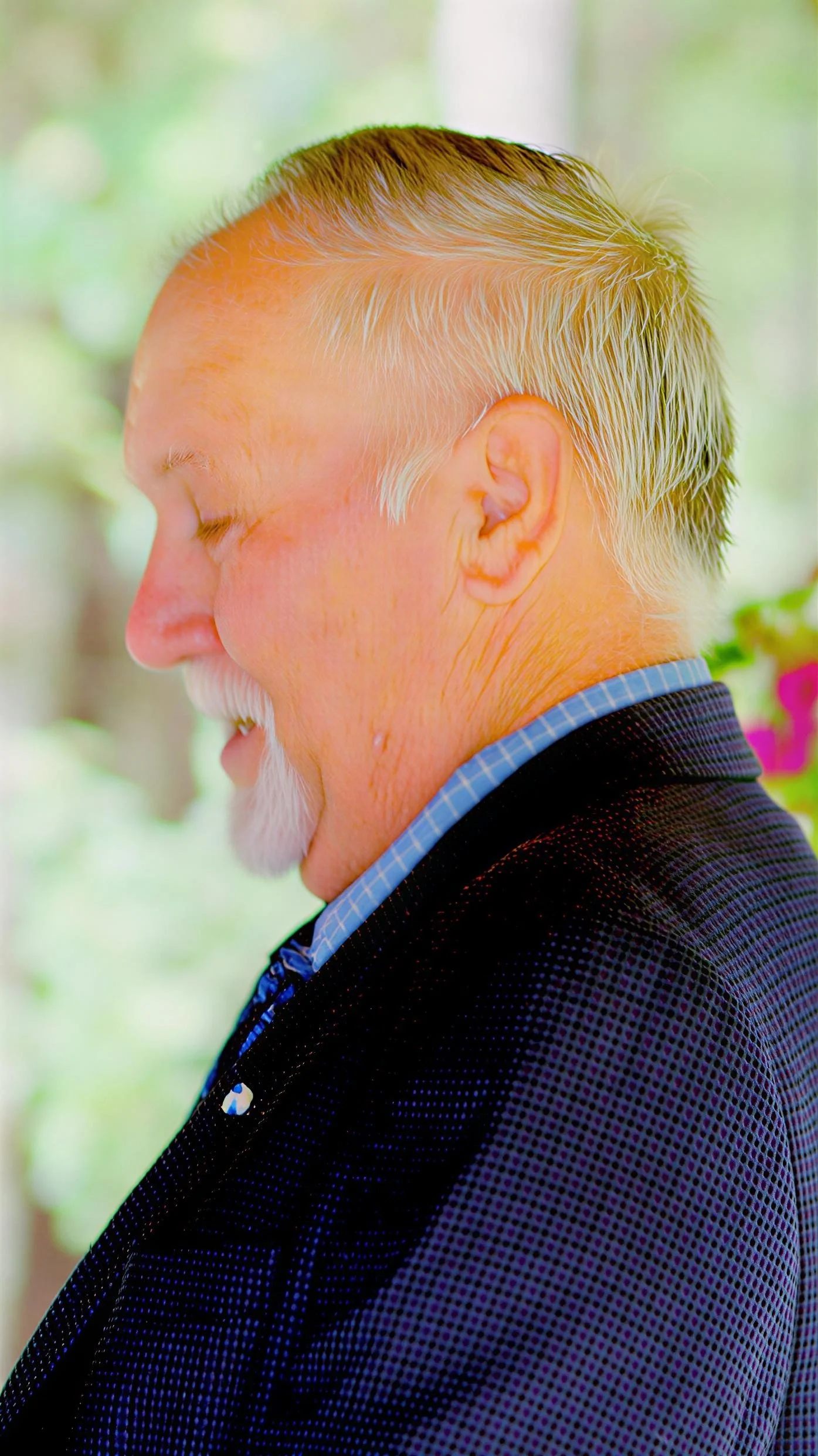 Side profile of an older man with a white goatee and blond hair, smiling with eyes closed, wearing a black suit jacket and blue checkered shirt, blurred green outdoor background.