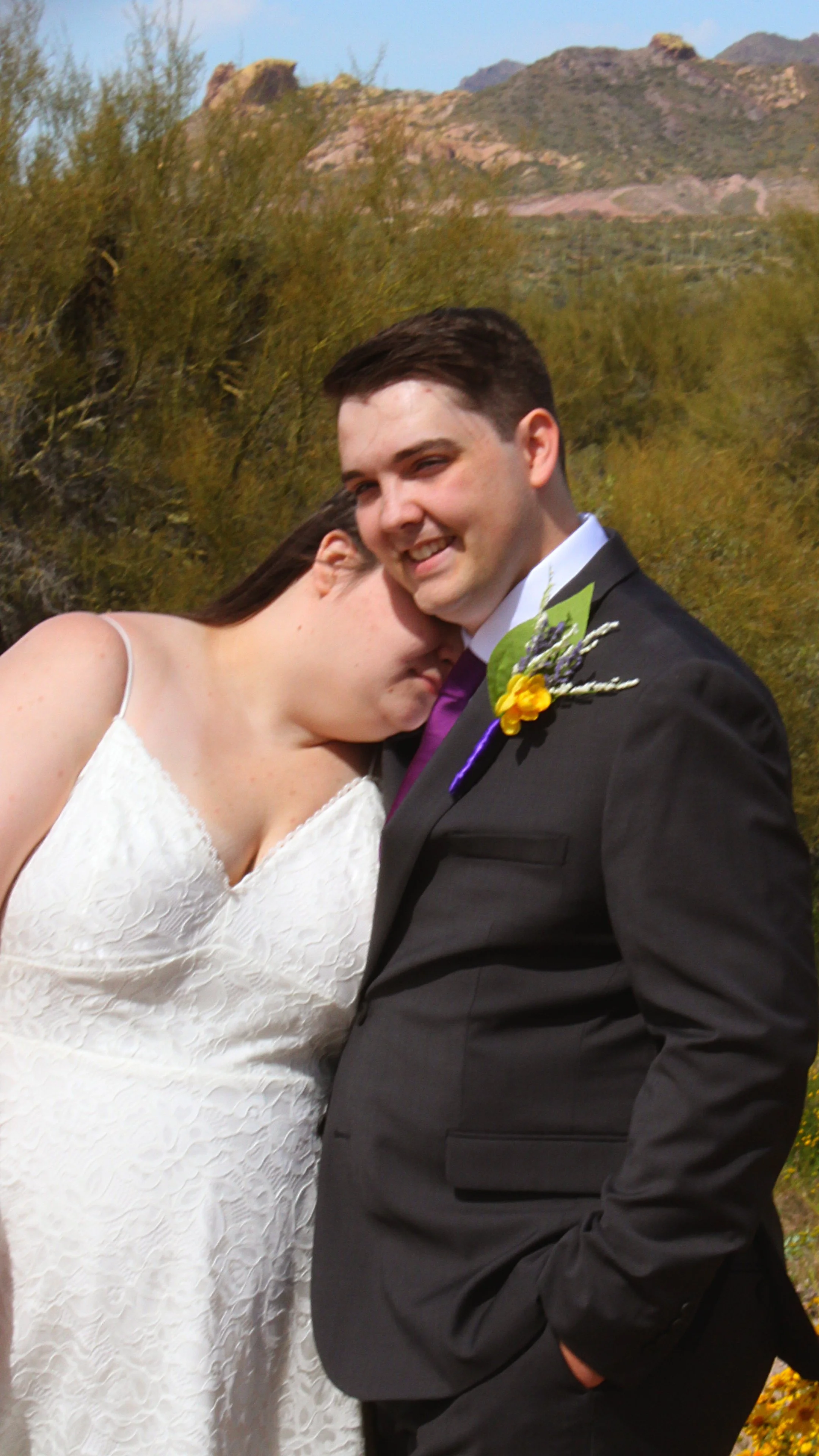 A smiling man in a suit and tie stands outdoors with a woman in a white dress leaning her head on his shoulder with closed eyes, in a natural setting with mountains and trees in the background.