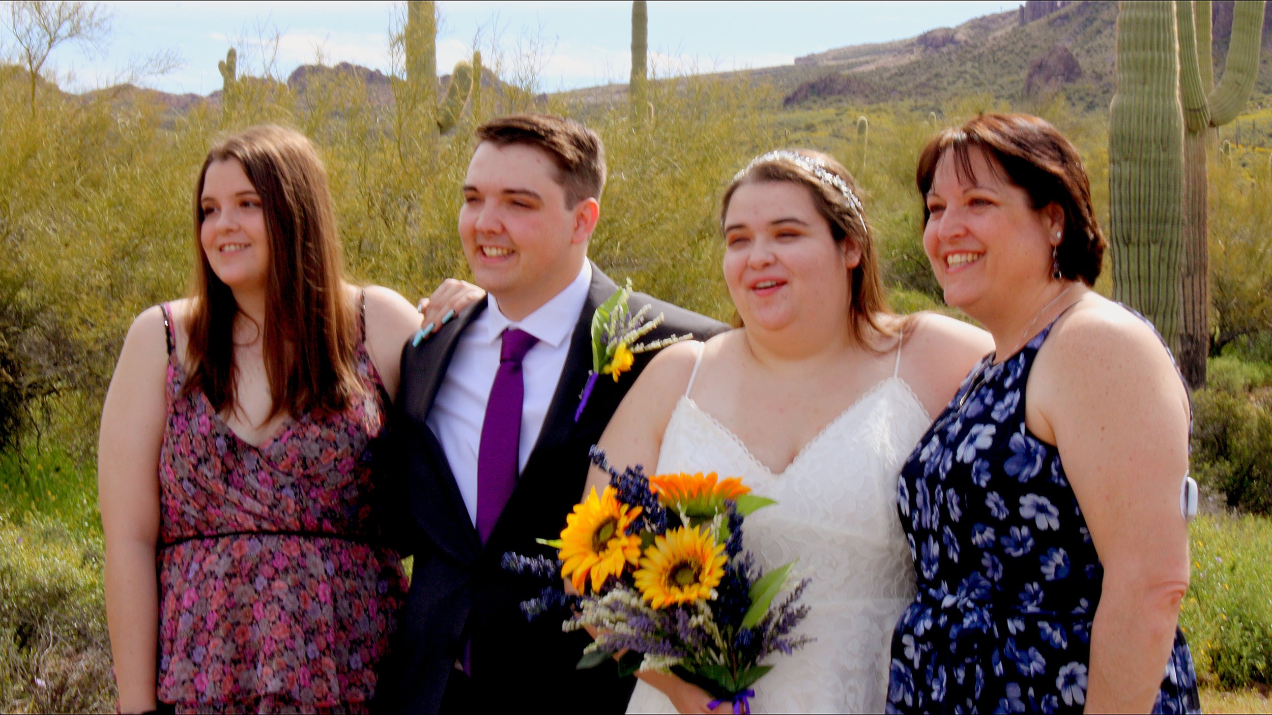 Four people at a wedding, standing outdoors in a desert landscape with cactus plants and mountains in the background. The man and woman in the middle are the newlyweds, with the woman holding a bouquet of sunflowers, and the others are family or frie