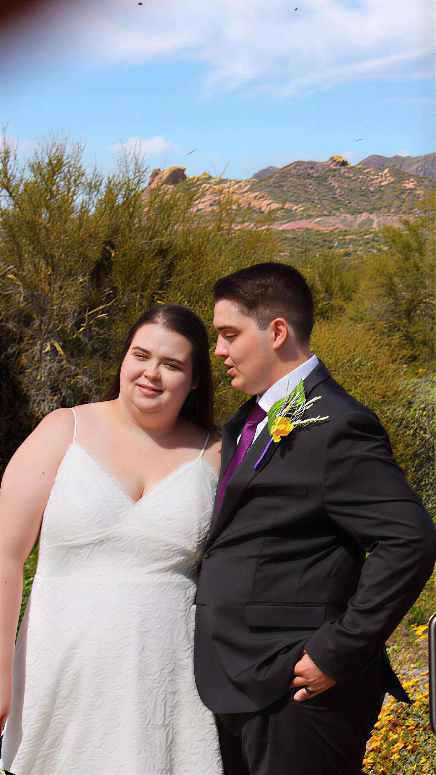 A newlywed couple standing outdoors with desert vegetation and mountains in the background. The bride is wearing a white dress, and the groom is in a black suit with a purple tie and a yellow boutonniere.