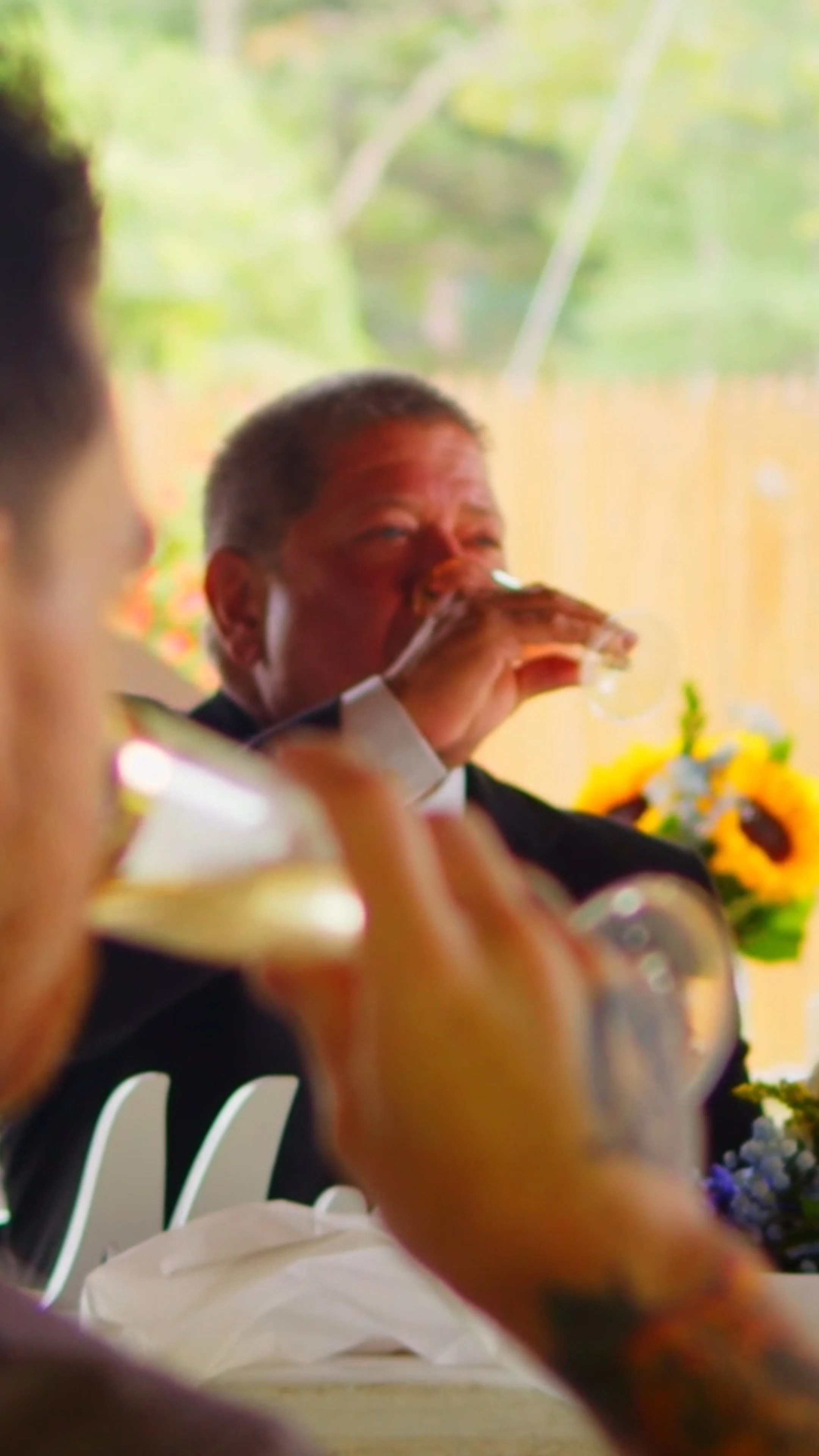 Person in formal attire drinking from a glass at a social gathering with bright outdoor scenery in the background.