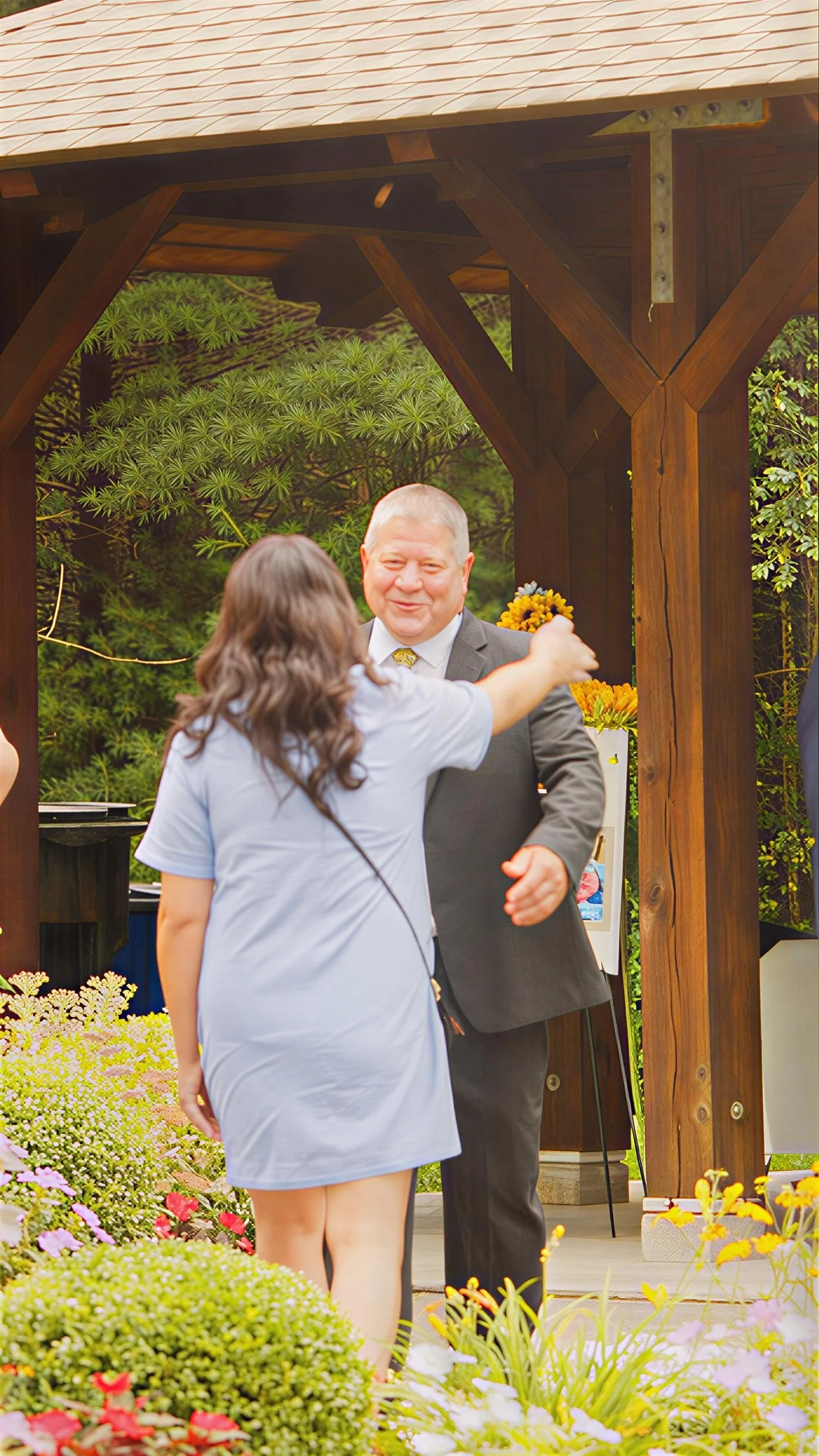A man in a suit smiling as a girl in a grey dress hugs him, standing on a garden patio with colorful flowers surrounding them.