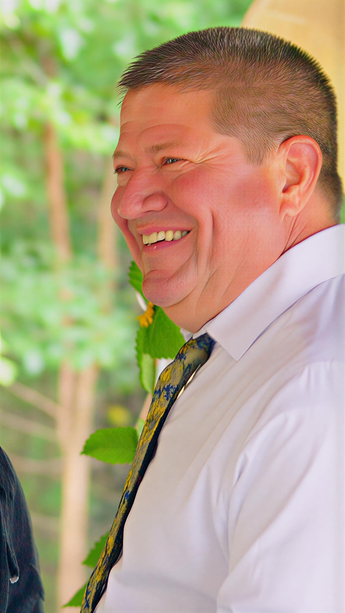 A smiling man wearing a white shirt and a colorful tie, with a background of green leaves and trees.